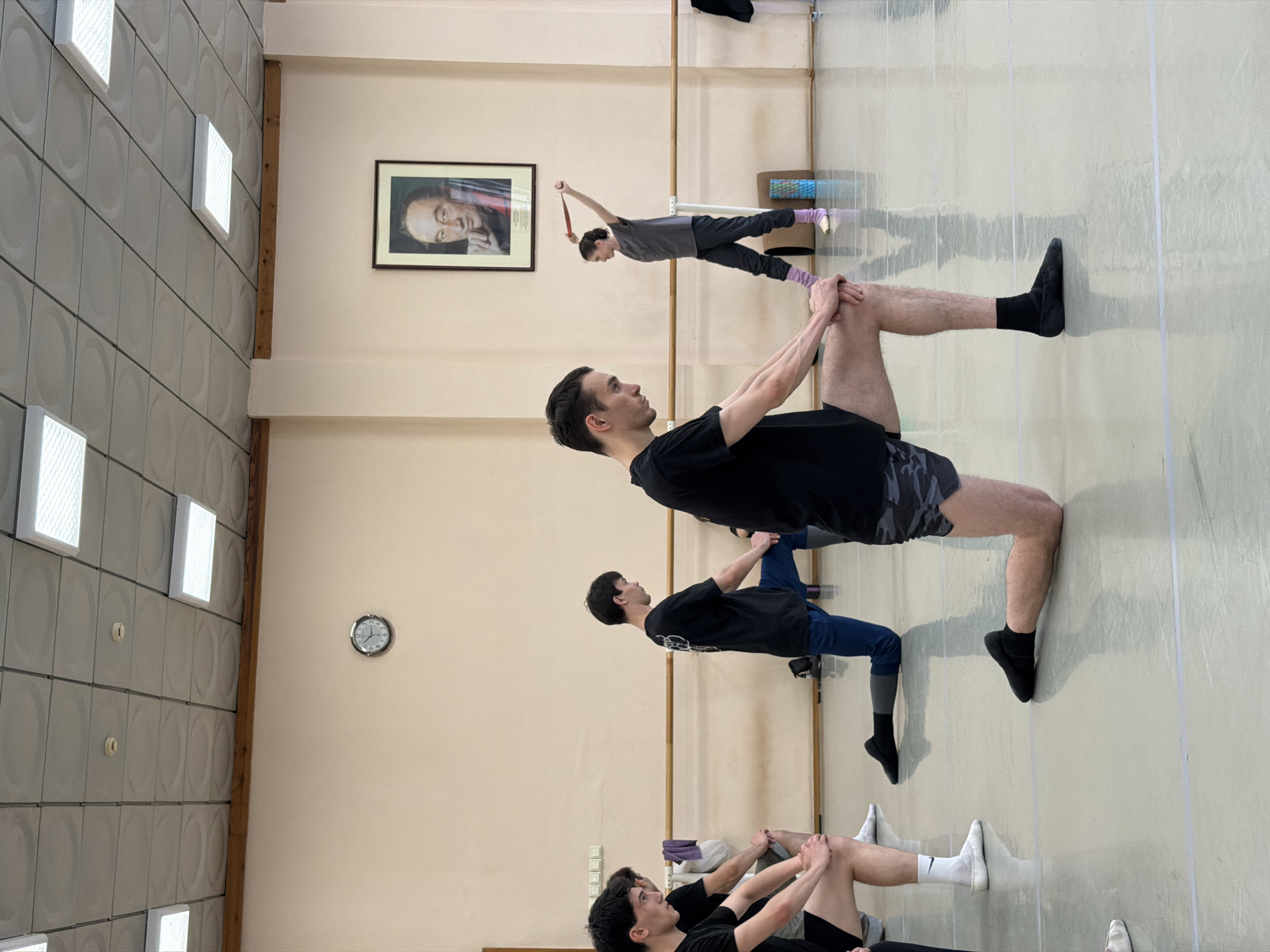 A group of male and female individuals are performing stretches in a dance studio. The room is spacious with a light-colored wall and a reflective floor. A barre runs along the wall, and a framed portrait of a man is displayed above it. The individuals are dressed in athletic wear, including t-shirts, shorts, and leggings. One person in the background is holding a resistance band. The overall scene suggests a dance class or rehearsal.