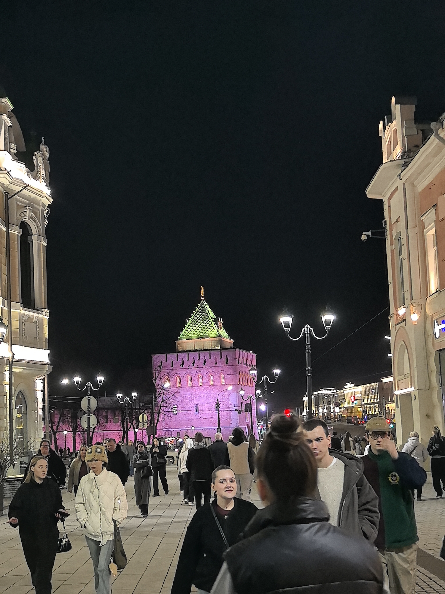 The image captures a street scene in Nizhny Novgorod, Russia, at night. A crowd of pedestrians are walking on a paved walkway lined by buildings on either side. The most prominent structure is a brightly lit tower with a green pyramid-shaped roof, likely part of the Nizhny Novgorod Kremlin. Streetlights illuminate the scene, and additional buildings are visible in the background. The crowd includes people of various ages and attire, suggesting an active urban environment.