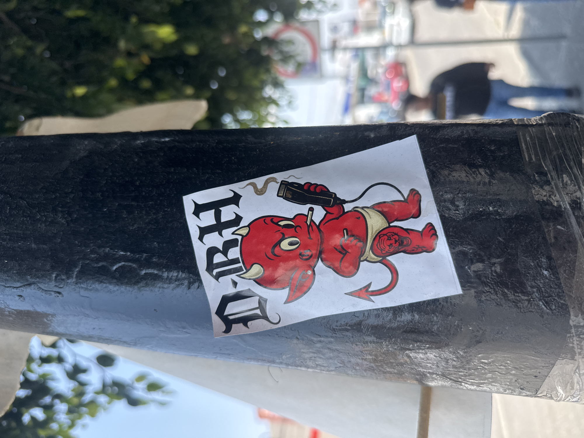 A sticker featuring a mischievous red devil baby is affixed to a textured, black surface, possibly a metal pole or railing. The devil baby, with prominent horns, yellow eyes, and a pointed tail, wears a beige diaper. It holds a black, corded device resembling an electric clipper or shaver, from which a smoky plume emanates. A thin, white object, possibly a cigarette or vape, is held between its lips. Tattoos are visible on the devil's limbs: a tiger-like face on its left arm and a star or swirl design on its right leg.
To the left of the devil, partially visible gothic-style text reads "BRAD". The sticker's colors are vibrant against the dark surface.
The setting appears to be outdoors in a bright, clear daytime environment. The background is softly blurred, showing green foliage from trees or bushes, distant urban structures, and indistinct figures of people, suggesting a street or public area in Puebla, Mexico. The overall impression is one of urban street art with a rebellious or cheeky undertone.