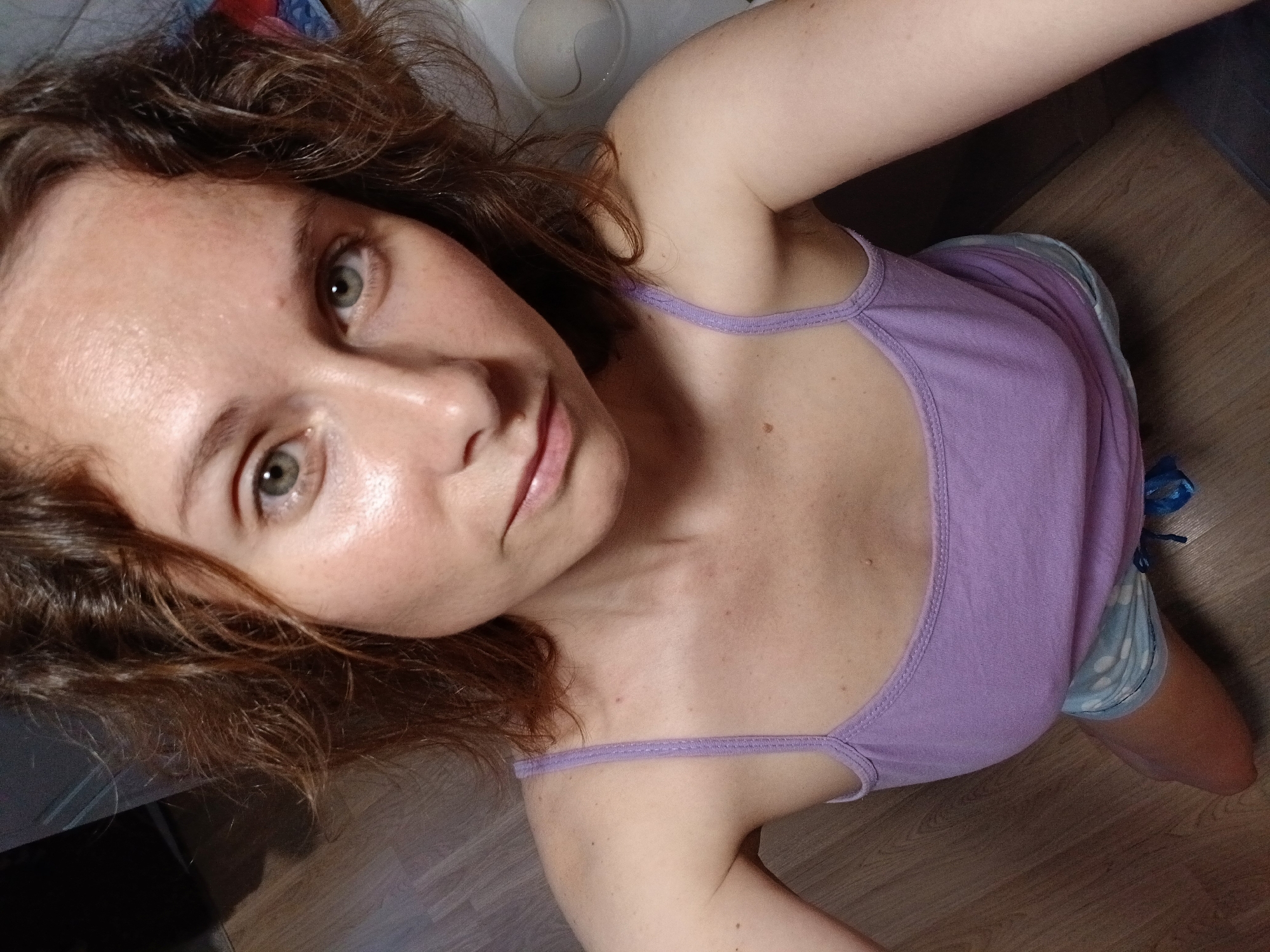 A high-angle, indoor, medium-shot shows a young Caucasian woman with curly brown hair and green eyes, looking directly at the camera. She is wearing a lavender tank top and light blue and white polka dot shorts. Her face is lightly powdered, and her lips are slightly parted. The background consists of a wooden floor and a white object on the right side of the frame. The scene appears to be a domestic setting, possibly a bedroom or living room. There is no indication of the specific location within Yekaterinburg, Russia.