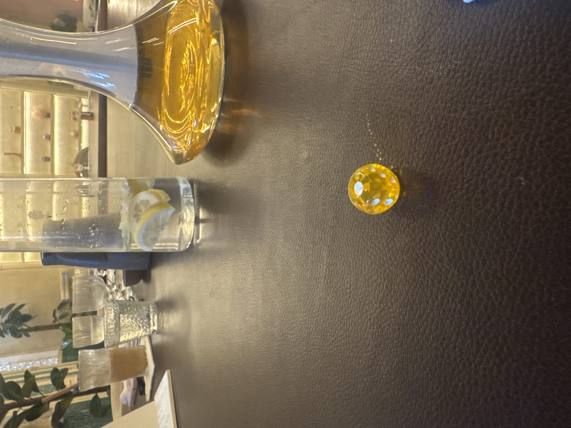 The image captures a table setting, seemingly indoors, with a dark, textured surface. On the left side, there is a glass pitcher containing a yellow liquid, possibly juice or water, along with a tall glass of water containing lemon slices. There are also a few small drinking glasses. A yellow candy is also on the table. Through the reflection in the glass pitcher, a glimpse of the interior can be seen, suggesting a room with ornate decor and greenery, suggesting a restaurant, hotel or home. The location is North Kuta, Indonesia.