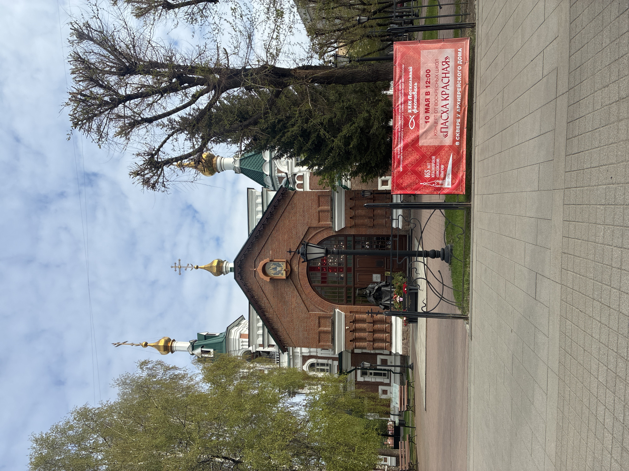 Here is a description of the image:
The image captures a daytime scene, focusing on a brick and white Orthodox church with golden domes topped with crosses. A red banner stands prominently in front of the church, advertising a "PASXA KPACHAYA" (Easter Red) concert on May 10th at 12:00, performed by Sunday schools. The banner also indicates it's the XXX Easter Festival and commemorates the 165th anniversary of the Krasnoyarsk (Yenisei) Eparchy.
The church is situated near a sidewalk paved with square gray tiles. Bare branches of trees frame the upper part of the image, with greenery partially obscuring the church's lower sections. A black iron fence separates the walkway from the church grounds. There are some street lights and a monument with flowers. The sky is partly cloudy. The scene appears to be in an urban setting, potentially a park or square.