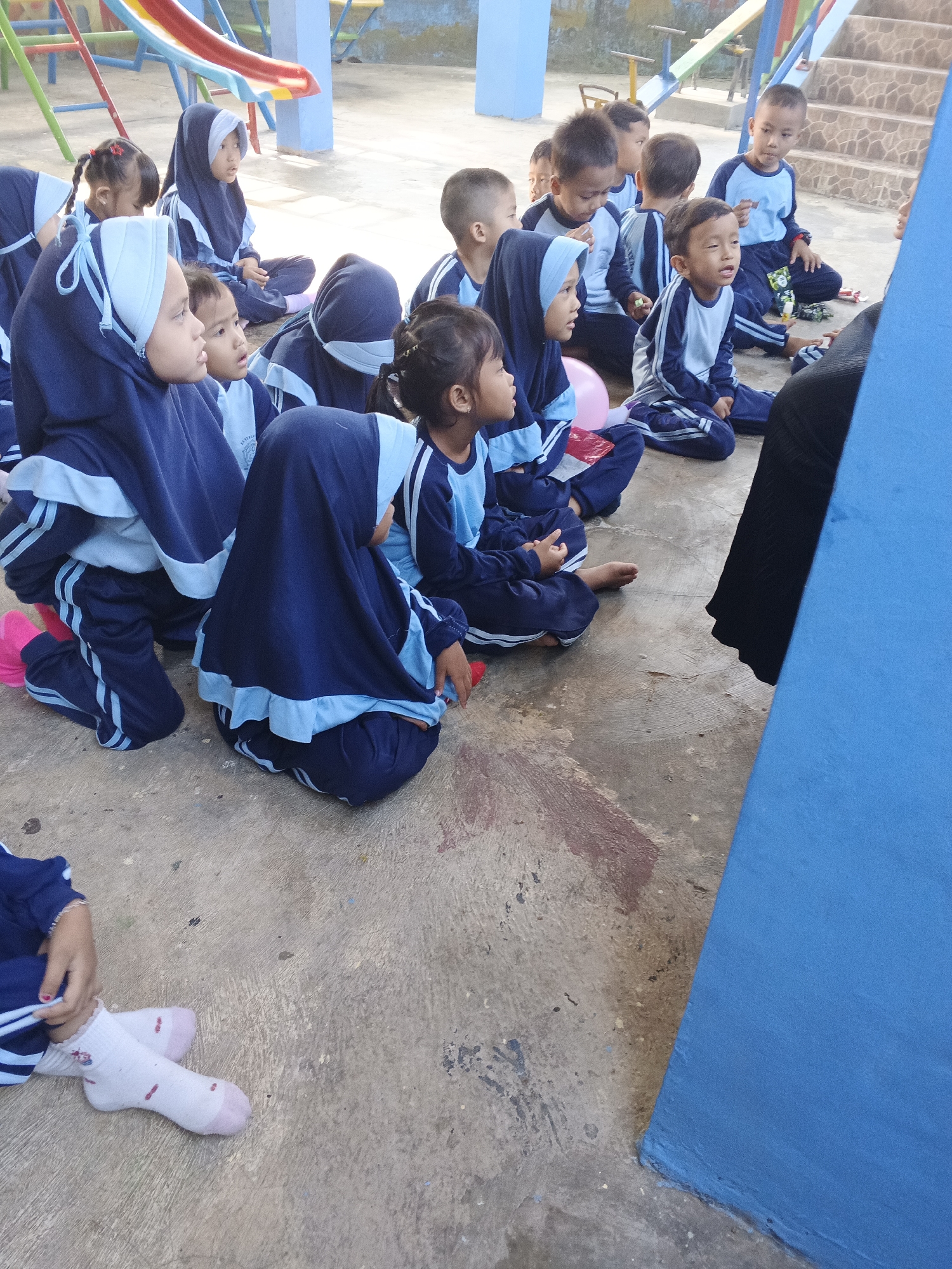 In Warungkiara, Indonesia, a group of children are seated on a concrete ground, facing a person out of frame. The children are wearing blue and white uniforms, with many of the girls also wearing hijabs. The scene appears to be an outdoor educational setting, possibly a schoolyard, with playground equipment visible in the background. The children are attentive, suggesting they are participating in an activity or lesson. The environment is somewhat worn, with paint marks on the concrete.