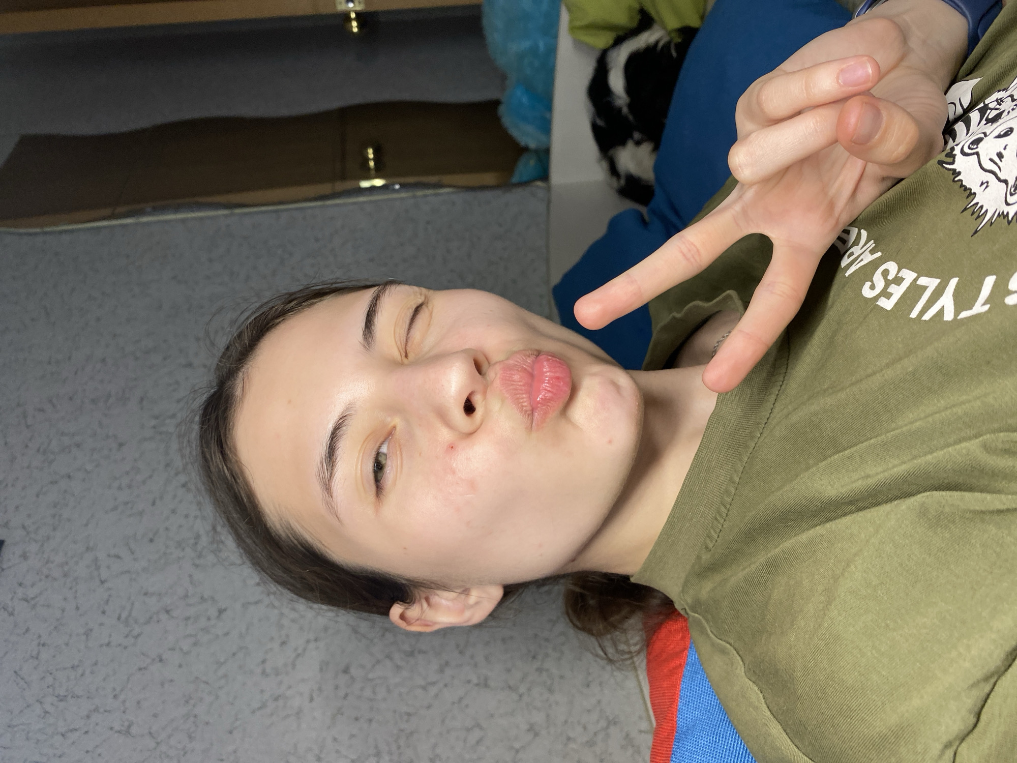 A young woman is facing the camera, winking one eye and making a kissy face with her lips. She is holding up a peace sign. She is wearing an olive green t-shirt with a white graphic design and possibly a patterned design on a blue, red, and white background underneath the green shirt. She has light skin and dark hair. The background is a grey surface with a hint of pattern, with objects that appear to be cabinets and fabric visible behind her. It is impossible to identify the location in Votkinsk, Russia, based on the image provided.