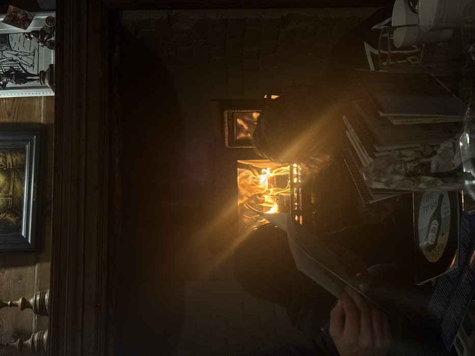 This image depicts an interior scene in an establishment located in Deal, United Kingdom, characterized by dim lighting and the warmth of an active fireplace.
In the mid-ground, a prominent fireplace contains a metal grate with bright orange flames from burning logs or coal, serving as the primary light source and casting strong lens flares across the scene. To the left of the fireplace, a person is seated, visible in silhouette from the chest down, holding an open document or menu with both hands. To the right of the fireplace, a second person is also seated, largely obscured and silhouetted, facing towards the fire.
Above the fireplace, a dark wooden mantlepiece supports decorative items. On the left, two turned wooden candlesticks are present. In the center, a framed portrait of a figure in historical attire is mounted. To the right, a smaller framed illustration or print, possibly depicting a figure or scene, stands beside a miniature figurine of a person riding a vintage motorcycle.
The foreground, particularly on the right, shows a cluttered table surface. Here, multiple spiral-bound booklets or menus are stacked upright. A small, light-colored decorative object or statue is visible. A dark circular object, possibly a coaster or placemat, displays partial white text including "TIME TO GO." Several white ceramic cups or mugs, some stacked, complete the table arrangement. The surrounding environment appears to have dark walls, possibly brickwork around the fireplace and wooden paneling above.