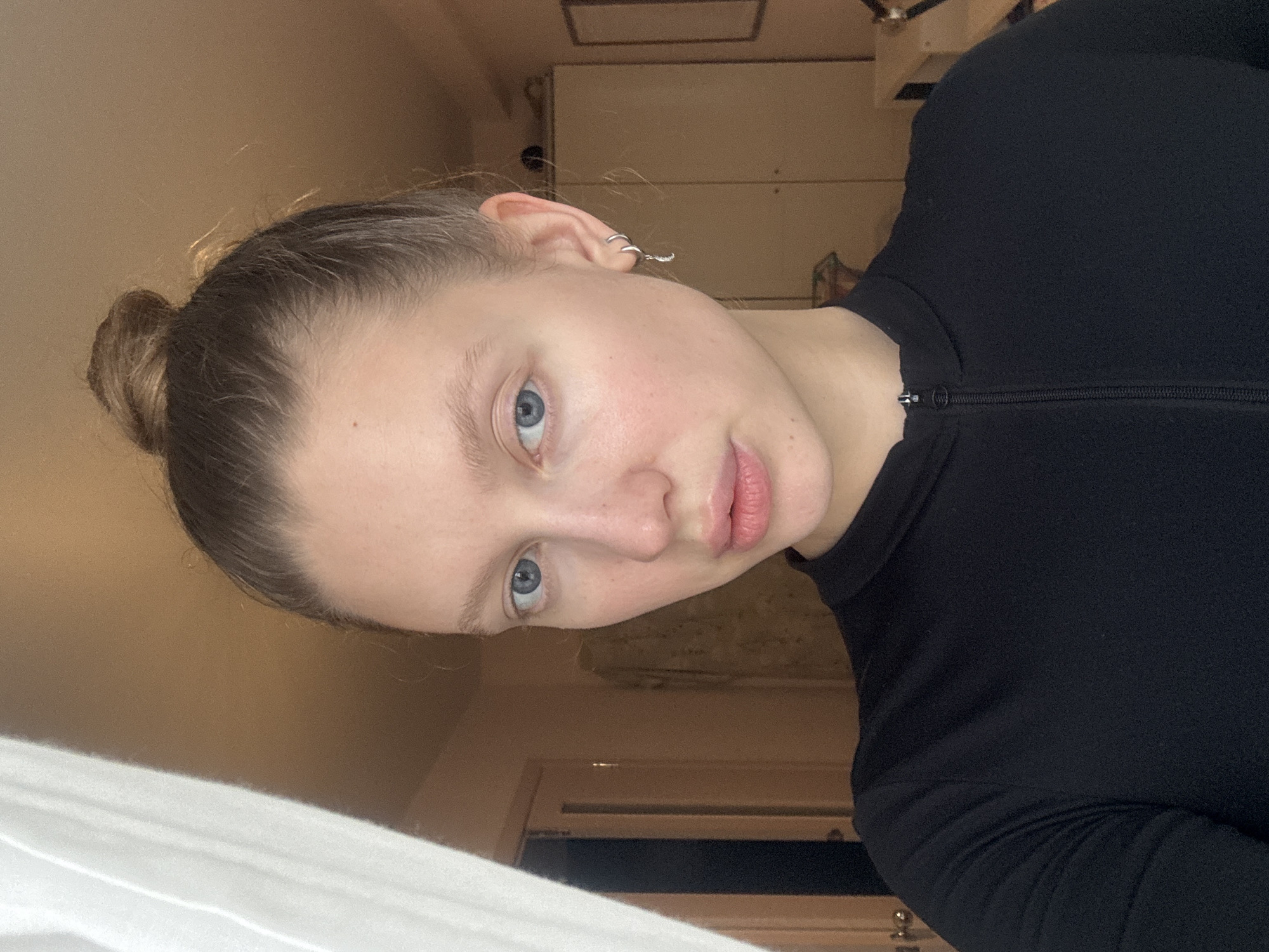 The image features a young adult female captured in a close-up, waist-up portrait within an indoor setting, understood to be in the city of Izhevsk, Russia. The subject has light skin, blue eyes, and light brown hair styled in a high, somewhat messy bun. Her expression is neutral, with a direct gaze towards the viewer, and her lips are slightly parted. She is wearing a black, long-sleeved top with a high collar and a visible zipper extending down the front. In her right ear (viewer's left), she wears two silver hoop earrings, one of which includes a small crescent moon charm. Her left ear (viewer's right) features a single silver hoop earring.
The background elements suggest a residential interior. On the far left, a vertical white element, likely a curtain or window frame, is visible. Adjacent to this, a light brown wall leads to a closed wooden door with a lighter pinkish-brown hue, featuring a darker vertical line to its left, possibly another door frame or shadow. The right side of the background shows a plain light brown wall, against which a large, light-colored, two-door wardrobe or cabinet is positioned. A rectangular mirror with a dark frame hangs on the wall to the right of the wardrobe. The overall lighting appears soft and even, highlighting the subject's face and providing general illumination to the room.