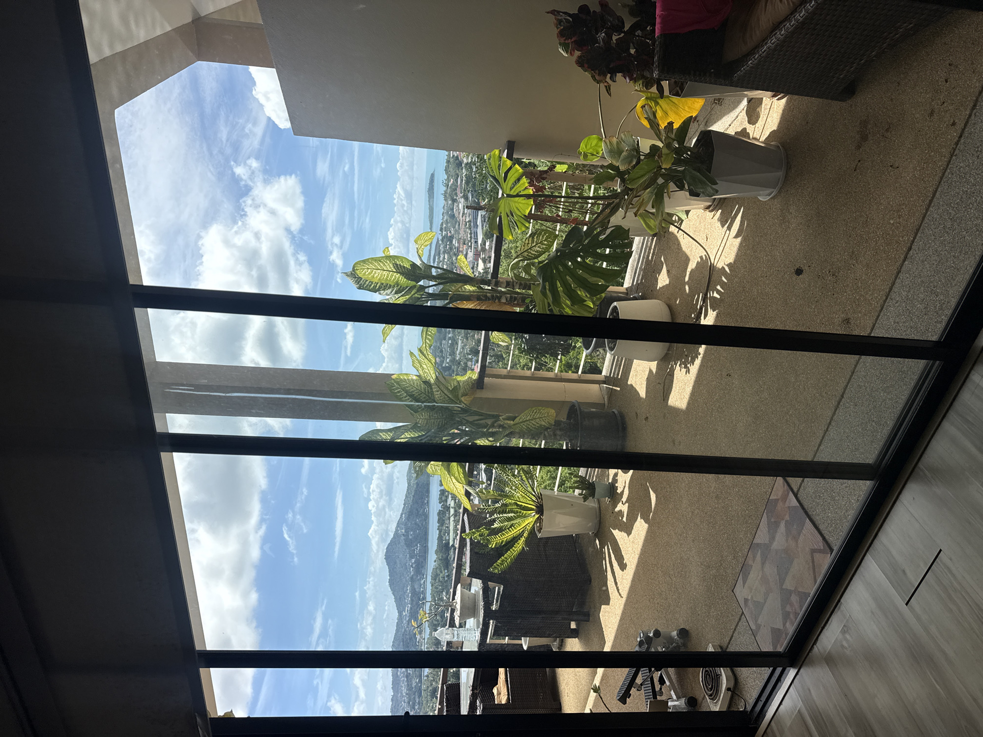 A wide-angle, exterior, eye-level shot captures a balcony with a view of a tropical landscape and ocean in Baan Chaweng, Thailand. The scene is viewed through a large, sliding glass door, with its dark framing partially obscuring the view. The balcony is furnished with wicker chairs and tables, some with cushions. Several potted plants, including a large monstera and a bird's nest fern, are arranged on the balcony floor. A water bottle rests on one of the tables. In the background, a sprawling city and lush green hills meet the turquoise ocean under a partly cloudy blue sky. Sunlight casts long shadows across the balcony floor. To the left of the balcony, a small exercise machine is visible.