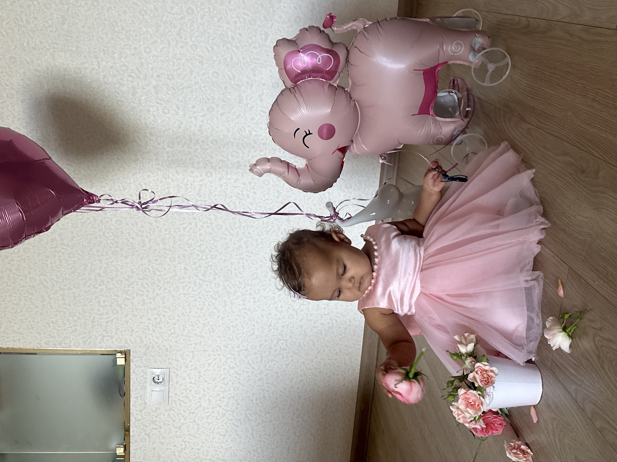 The image depicts a baby girl dressed in a pink tutu and dress, sitting on a wooden floor indoors. She is holding a pink rose and looking down at a small bucket filled with pink roses and greenery. To her right is a large pink elephant balloon on wheels, and a smaller gray elephant figurine. A pink heart-shaped balloon is visible in the upper right corner of the image. The background features a textured light-colored wall with a power outlet and a framed mirror above it. This scene appears to be a domestic setting, possibly a child's room or a living area, not specifically indicative of Petrozavodsk, Russia. The lighting suggests an indoor environment, with a soft natural light source, casting subtle shadows. The overall ambiance is festive and celebratory, likely for a birthday or special occasion.