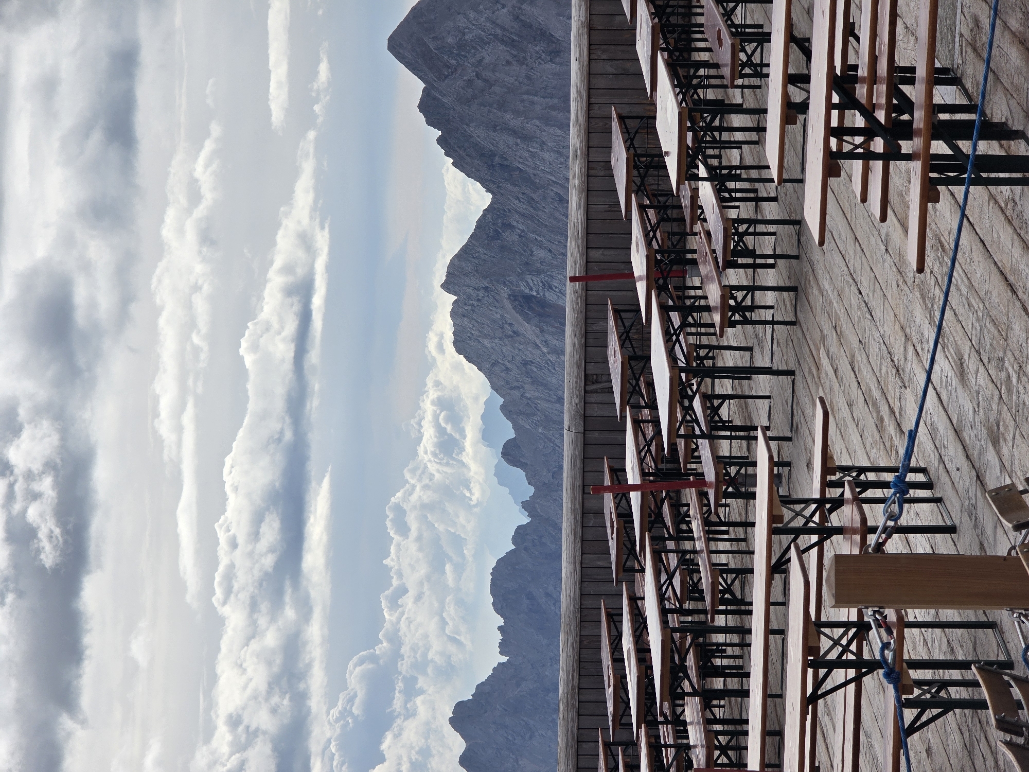 The image depicts an outdoor seating area with numerous wooden picnic tables and benches set up on a wooden deck. The tables are arranged in rows and appear to be empty. In the background, a wooden railing separates the seating area from a mountainous landscape. The mountains are rugged and appear to be covered in sparse vegetation or rock. The sky is overcast with dramatic clouds. This scene is likely from the Zugspitze, Germany's highest peak, located near Garmisch-Partenkirchen. The weather suggests it may be a cooler day, possibly autumn or spring, given the cloud cover and the absence of people.