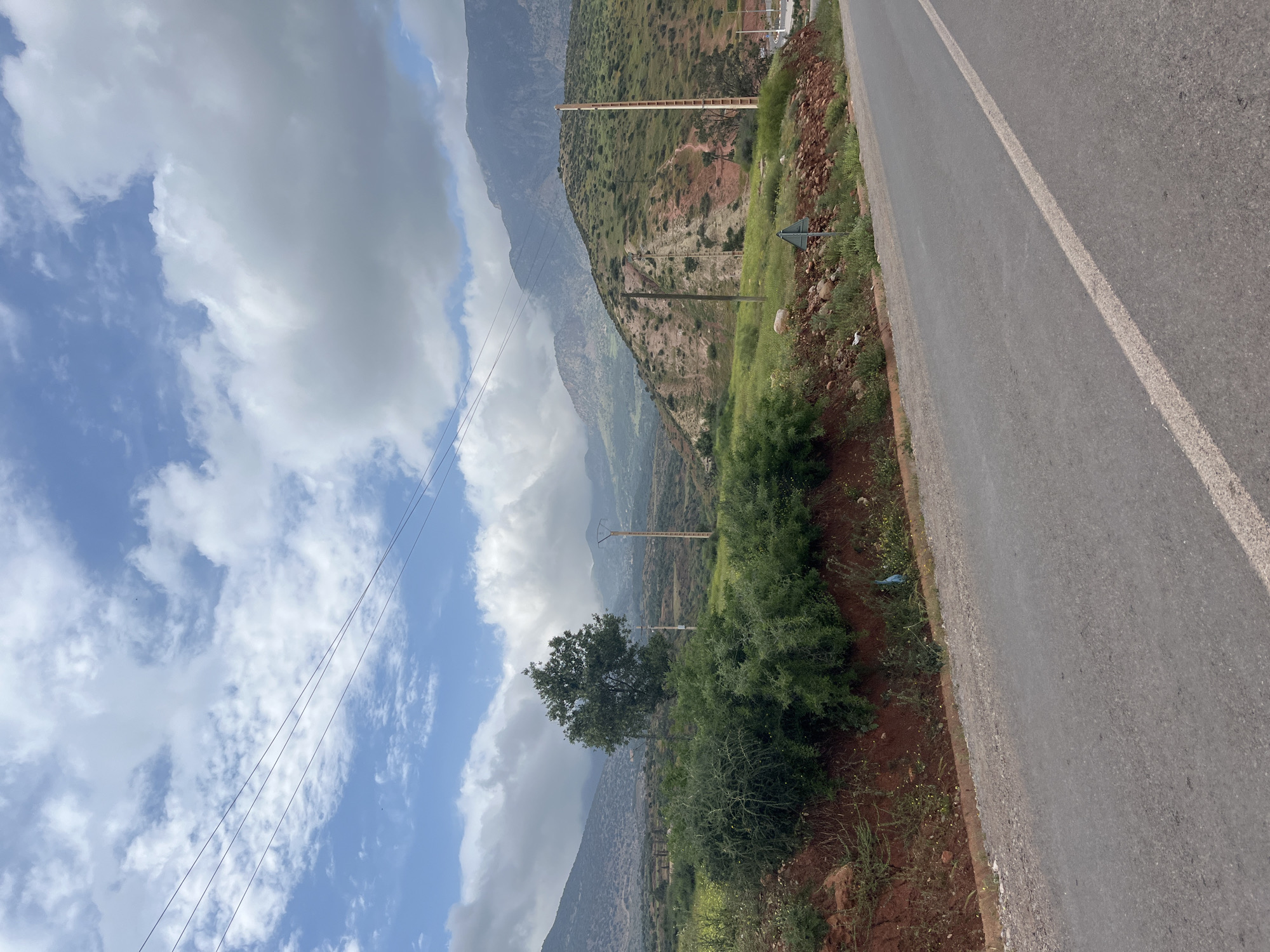 The image displays a picturesque mountainous landscape viewed from the side of a road. A grey asphalt road runs along the right side of the frame, marked by a clear white line. Adjacent to the road, the ground is a mix of reddish-brown earth and green vegetation, including sparse grass and small shrubs.
To the left of the road and in the mid-ground, a prominent green tree stands amidst denser bushes and vegetation. Beyond this foliage, a series of mountains dominate the left and background of the scene. These mountains exhibit varying textures and colors, with areas of lush green vegetation transitioning to more barren, reddish-brown rock formations. Some distant peaks are partially obscured by clouds or atmospheric haze. Utility poles and power lines traverse the scene horizontally, extending across the mid-ground and background, suggesting the presence of infrastructure in this remote area. A small, triangular road sign is partially visible near the road on the right.
The sky above is a vibrant blue, adorned with an array of white and light grey clouds, indicating a partly cloudy day. The bright lighting suggests it is daytime, likely late morning or early afternoon. The overall impression is one of natural beauty and rugged terrain, characteristic of a mountainous region like Ouaouizert, Morocco. There are no people visible, and no specific activities are taking place. No discernible text is visible on any objects within the image.
