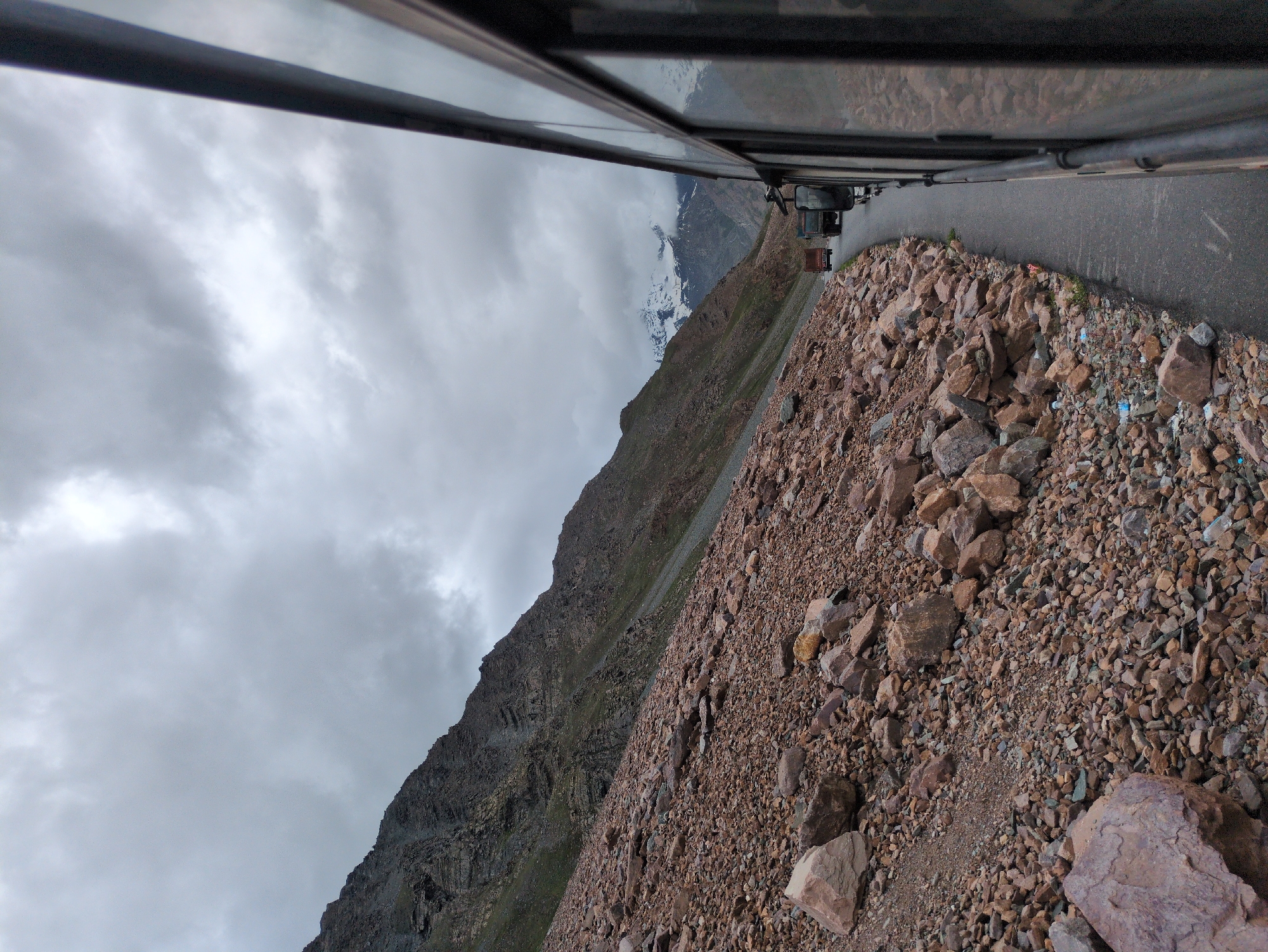 This is a photograph taken from inside a moving vehicle, looking out at a mountain road. The perspective is from the right side of the frame, with the side of the vehicle and its windows dominating the foreground and right edge. The road curves to the left, leading into a valley. On the left side of the road, a steep, rocky mountainside rises, covered in loose stones and scree. Some sparse green vegetation is visible on the higher slopes.
In the distance, the road continues into a mountain pass, where two trucks are visible. Further beyond the trucks, snow-capped mountains rise against a cloudy, overcast sky. The sky is filled with dramatic, grey clouds, suggesting inclement weather. The overall impression is one of a remote, rugged mountain landscape. This scene is located on the Baralacha Pass road, a high mountain pass in the Zanskar range, on the Leh-Manali Highway, in the region of Zing Zing Bar, India.