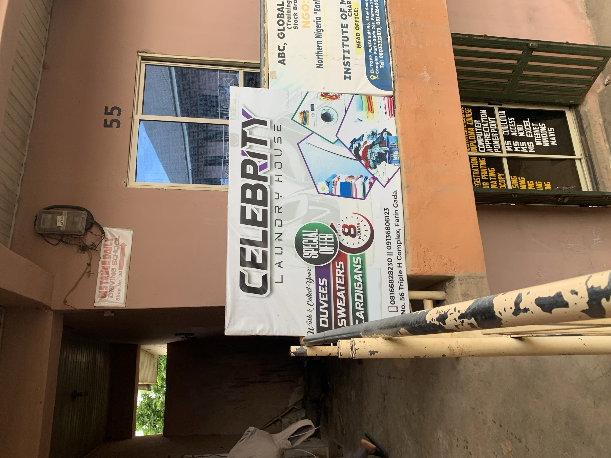 The image depicts the exterior of a building in Jos, Nigeria. A large banner for "CELEBRITY LAUNDRY HOUSE" is prominently displayed, advertising services like washing, duvets, sweaters, and cardigans with a special offer of service in 8 hours. Adjacent to this banner is a smaller sign for "ABC, GLOBAL (Training & Stock Broker)" and "INSTITUTE OF MANAGEMENT AND CHARTERED SECRETARIES." The building appears to be a commercial establishment, with a shop number "55" visible above a window. The scene also includes a pedestrian walkway with a yellow railing, and a doorway leading to an interior space displaying signs for computer courses and services such as "MS COREL DRAW," "MS ACCESS," and "MS EXCEL." A small sign for "DISTANCE DAILY DRIVING SCHOOL" is also visible on the upper portion of the building.