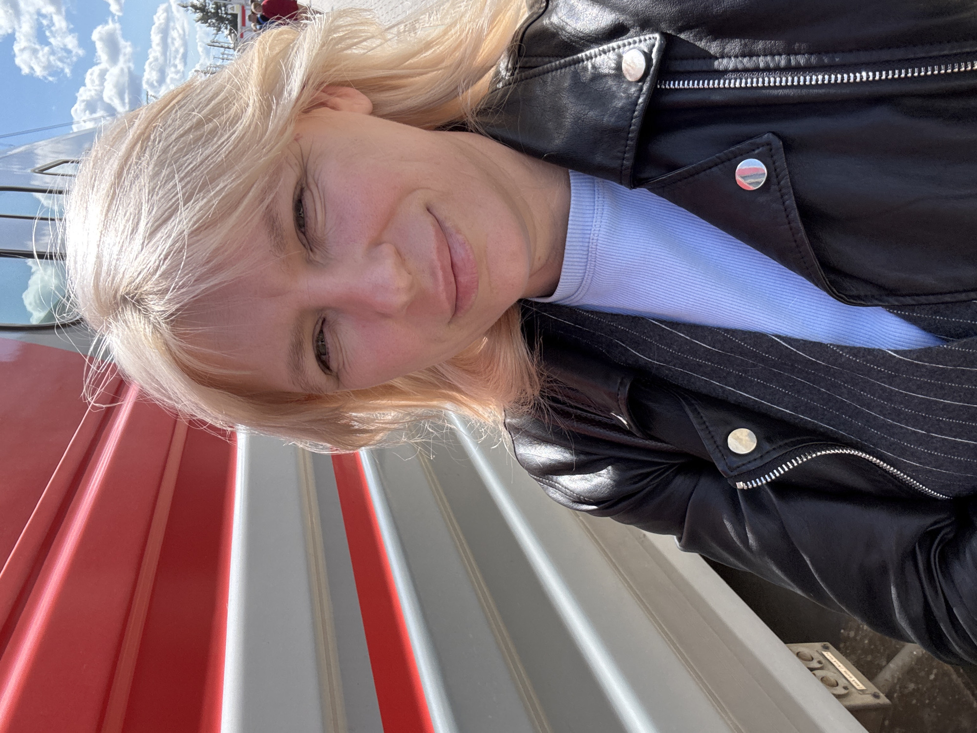 In the city of Rossosh, Russia, a woman with blonde hair, wearing a black leather jacket over a blue ribbed shirt, takes a selfie in front of a train car. The train car has red and grey horizontal stripes. The woman is looking at the camera with a slight smile. The background shows a bright, sunny sky with some clouds.