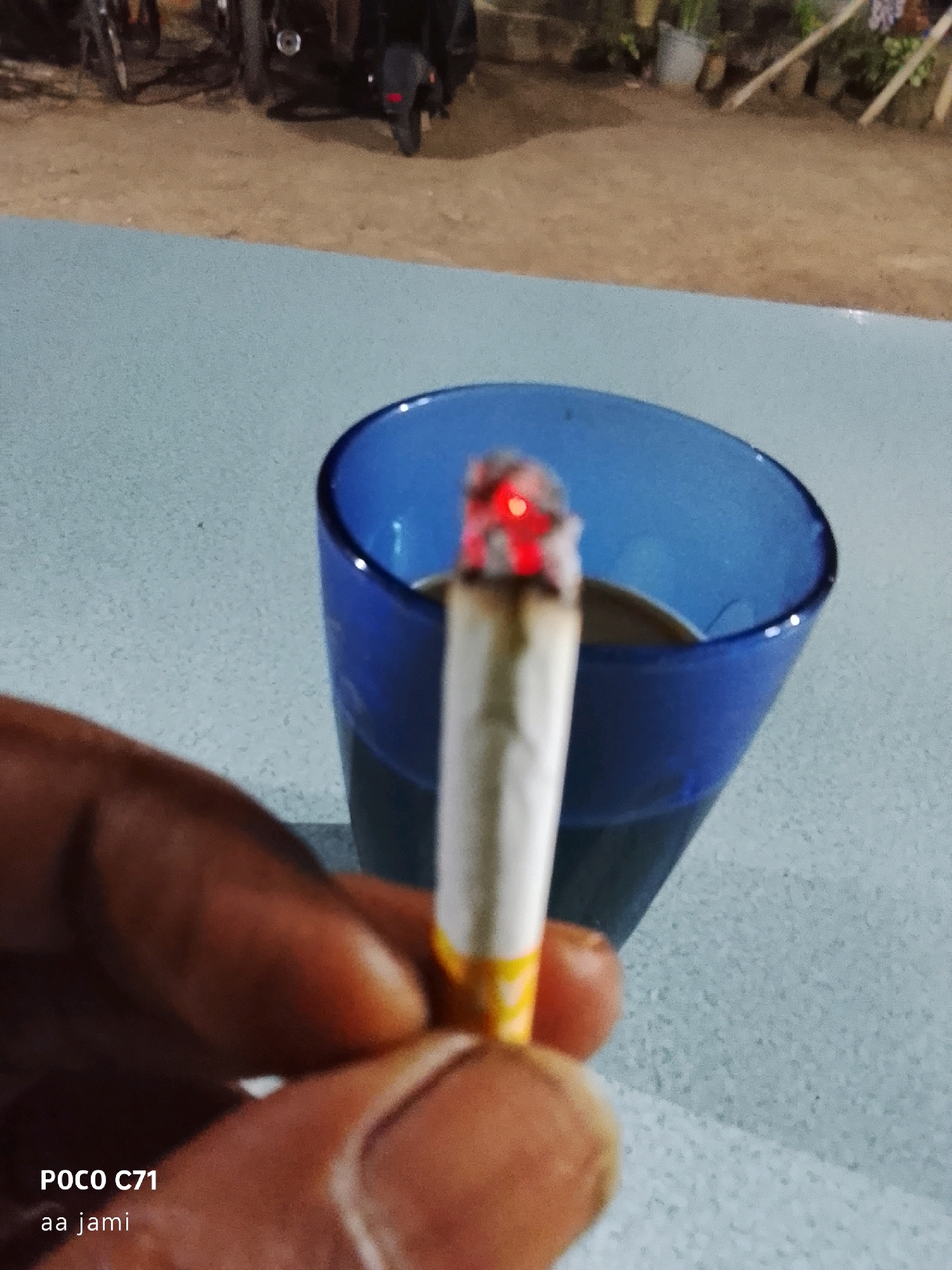 The image is a close-up, slightly blurry shot focused on a hand holding a lit cigarette. The cigarette, with a glowing red tip, is positioned in front of a blue glass containing a dark liquid, likely coffee. The background shows a dusty, outdoor area with what appears to be parked motorcycles and some greenery. The text "POCO C71" and "aa jami" are visible in the lower left corner, suggesting this is a photograph taken with a Poco C71 phone and possibly captioned by someone named Jami. The scene evokes a casual, possibly outdoor setting.