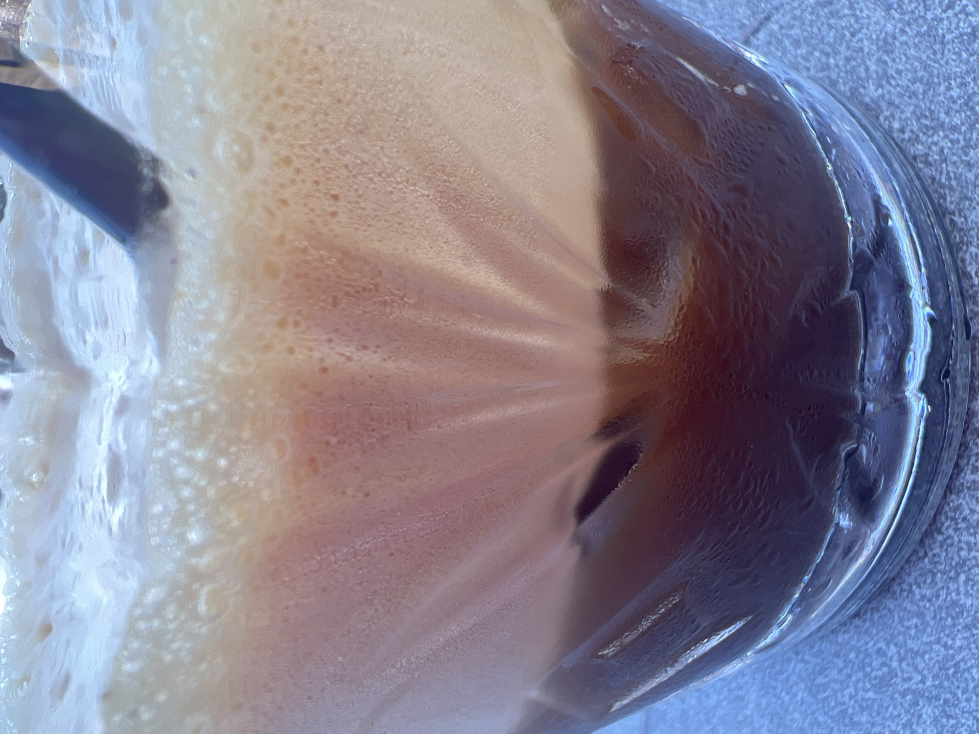 This is a close-up, vertical shot of a glass of dark liquid with a foamy head, likely a beverage like iced coffee or a soda. The glass is textured with vertical ridges that refract the light. The liquid at the bottom is a deep brown, while the foam at the top is lighter, creamy white with hints of pink and orange. There are small bubbles throughout the foam. A dark straw is visible protruding from the foam. The glass appears to be sitting on a textured surface, possibly a table, that is light gray with a speckled pattern. The lighting suggests an outdoor setting, possibly in the city of Eretria, Greece, on a bright day, as indicated by the condensation on the glass. No people are visible in the image.