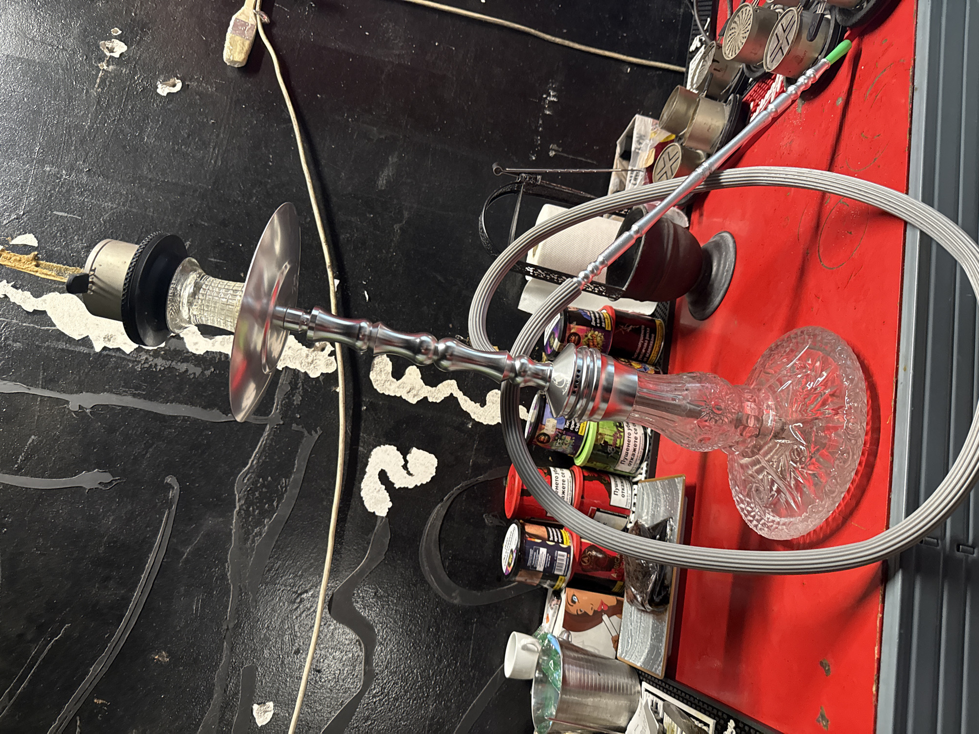 This image features a hookah, prominently displayed against a cluttered backdrop of various items. The hookah is silver, with a clear, patterned glass base. Its hose is gray and loops around a cluster of small containers, possibly tobacco flavorings, with labels in Cyrillic script suggesting they are from a Bulgarian or Slavic-speaking region.
The surroundings include a black, textured surface reminiscent of a tabletop, possibly damaged, showing white paint chips. This contrasts with a red surface that may be a section of a countertop or shelving unit, where the hookah base and flavorings are situated. A selection of foil disks for the hookah, smoking accessories, and a brush are also present.
Without further context, it's not possible to confidently pinpoint a location in Plovdiv, Bulgaria. The scene appears to be indoors, likely in a commercial or recreational establishment related to hookah use.