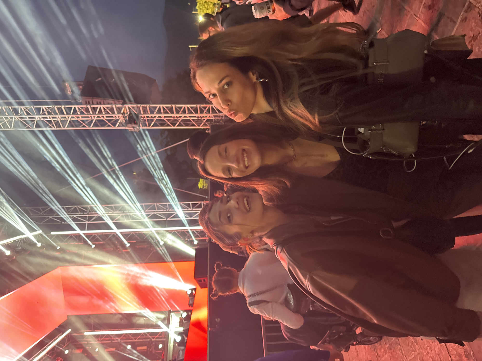 Three young women pose for a photo at an outdoor event, possibly a concert or festival, in Budva, Montenegro. A stage with white spotlights and red lighting is in the background. The top woman has long brown hair, a black leather jacket, and is making a pouting face. The middle woman is smiling and wearing a dark top. The bottom woman is wearing a brown jacket. Other people are visible in the background.