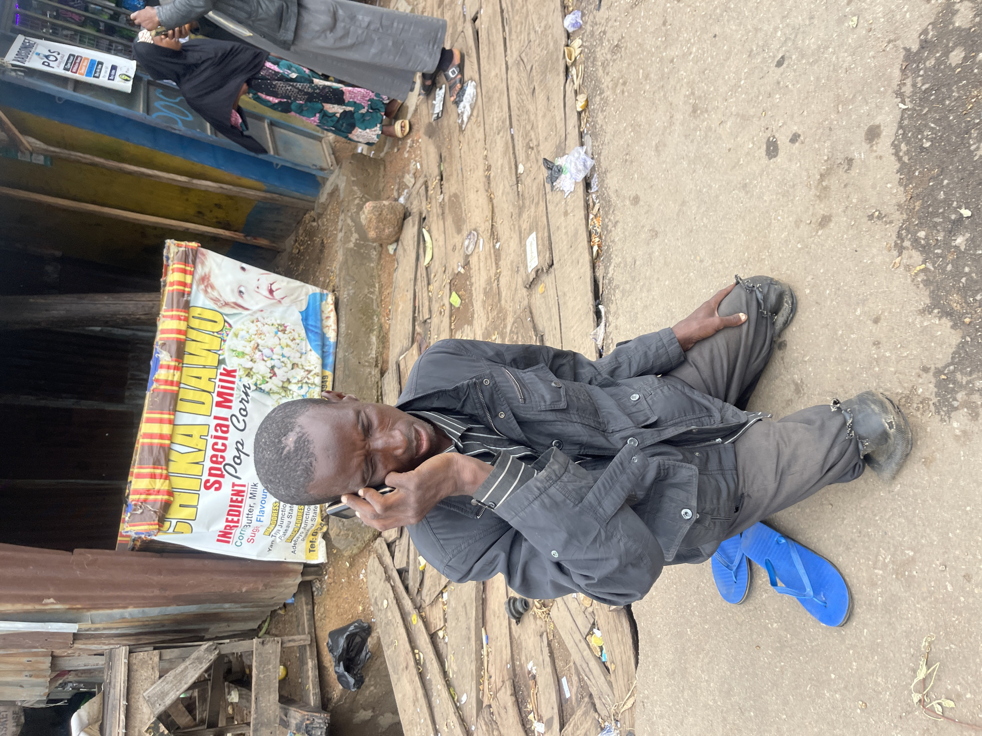 A Black male kneels on a concrete surface, speaking on a black mobile phone held to his right ear. He is wearing a dark gray jacket over a striped shirt and dark gray pants. His left hand rests on his left knee, and he is barefoot, with a pair of bright blue flip-flops lying next to his left leg.
Behind him, a stall features a banner advertising "SHIKA DAWO Special Milk Pop Corn." The stall's structure is made of corrugated metal and wood. Scattered wooden planks and debris are visible on the ground. To the right, two other individuals are partially visible. One, wearing a black hijab and a patterned dress, stands near a blue and yellow storefront. Another, wearing a gray garment, stands behind the first.
The environment appears to be an outdoor market or street scene in Jos, Nigeria, characterized by its makeshift stalls and the presence of everyday commerce. The ground is a mix of concrete and uneven wooden planks, with scattered trash. The overall impression is one of a busy, possibly dusty, urban marketplace.