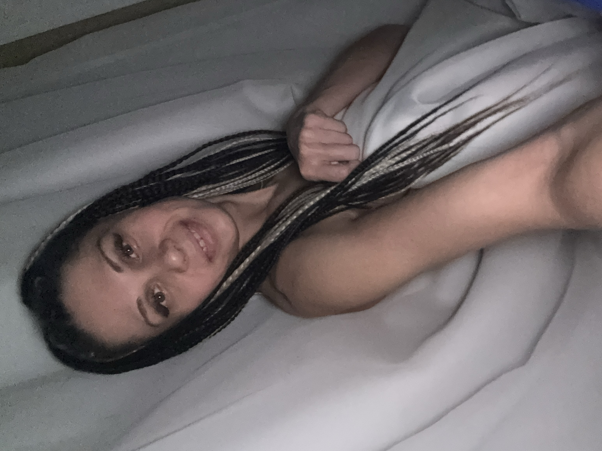 A person with dark hair braided with blonde strands lies in a white bed. The person is smiling and has their fist clenched near their chest. The location is unknown.