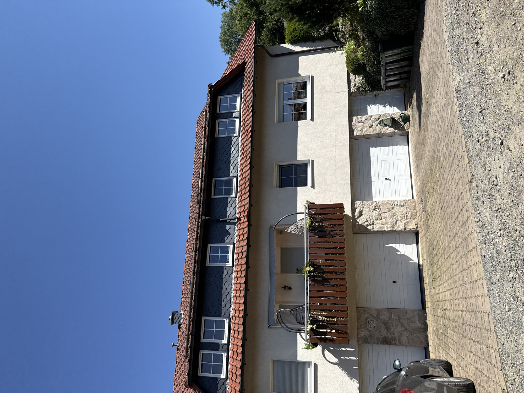 The image shows a two-story house with a red tiled roof and dark gray slate siding on the upper level under the roofline. The house has several windows with white frames and shutters. A wooden balcony with a brown railing is visible on the second floor, furnished with hanging chairs and planters. Below the balcony, there are three white garage doors on the ground floor, two of which are large and one is smaller. The area in front of the garages is paved with interlocking bricks. A dark-colored car is parked to the left of the garages. To the right of the garages, there are several green trash bins and dense vegetation. The house appears to be located in a residential area, possibly a suburb, of Bad Lauterberg im Harz, Germany, given the architectural style and the presence of the garage doors. The sky is clear and blue, indicating a sunny day.