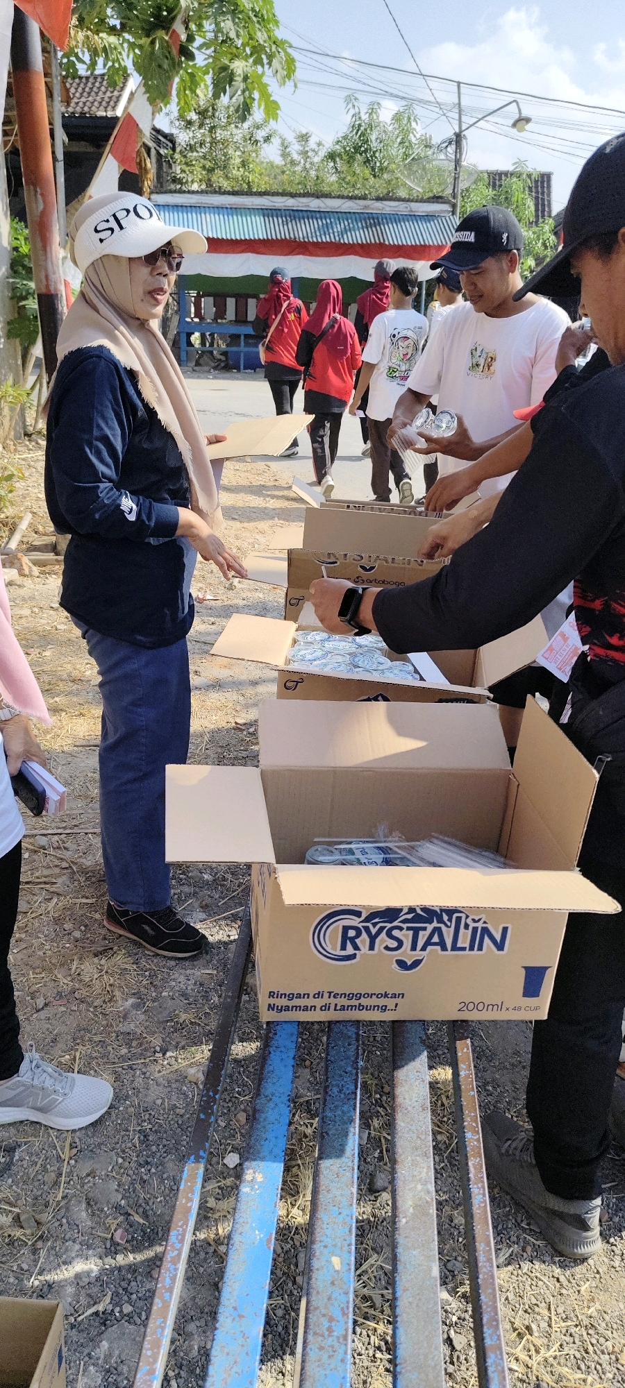 A group of people are gathered outdoors, possibly distributing goods. In the foreground, a woman wearing a beige hijab and a dark blue long-sleeved shirt stands to the left, holding a piece of cardboard. She is wearing blue jeans and dark sneakers. To her right, a person wearing a black long-sleeved shirt and dark pants is handing out clear plastic cups from a cardboard box labeled "Crystalin." Another person wearing a white t-shirt with graphics is visible behind the box. In the background, several other individuals, some wearing red shirts and hijabs, are walking away. The ground is covered in dry grass and gravel. A metal bench, painted blue and brown, runs horizontally across the lower portion of the image. Behind the people, a building with a red and white striped awning is visible, along with trees and a clear sky. The scene appears to be in a rural or semi-urban area, possibly within the city of Sulang, Indonesia, given the Indonesian text on the box.