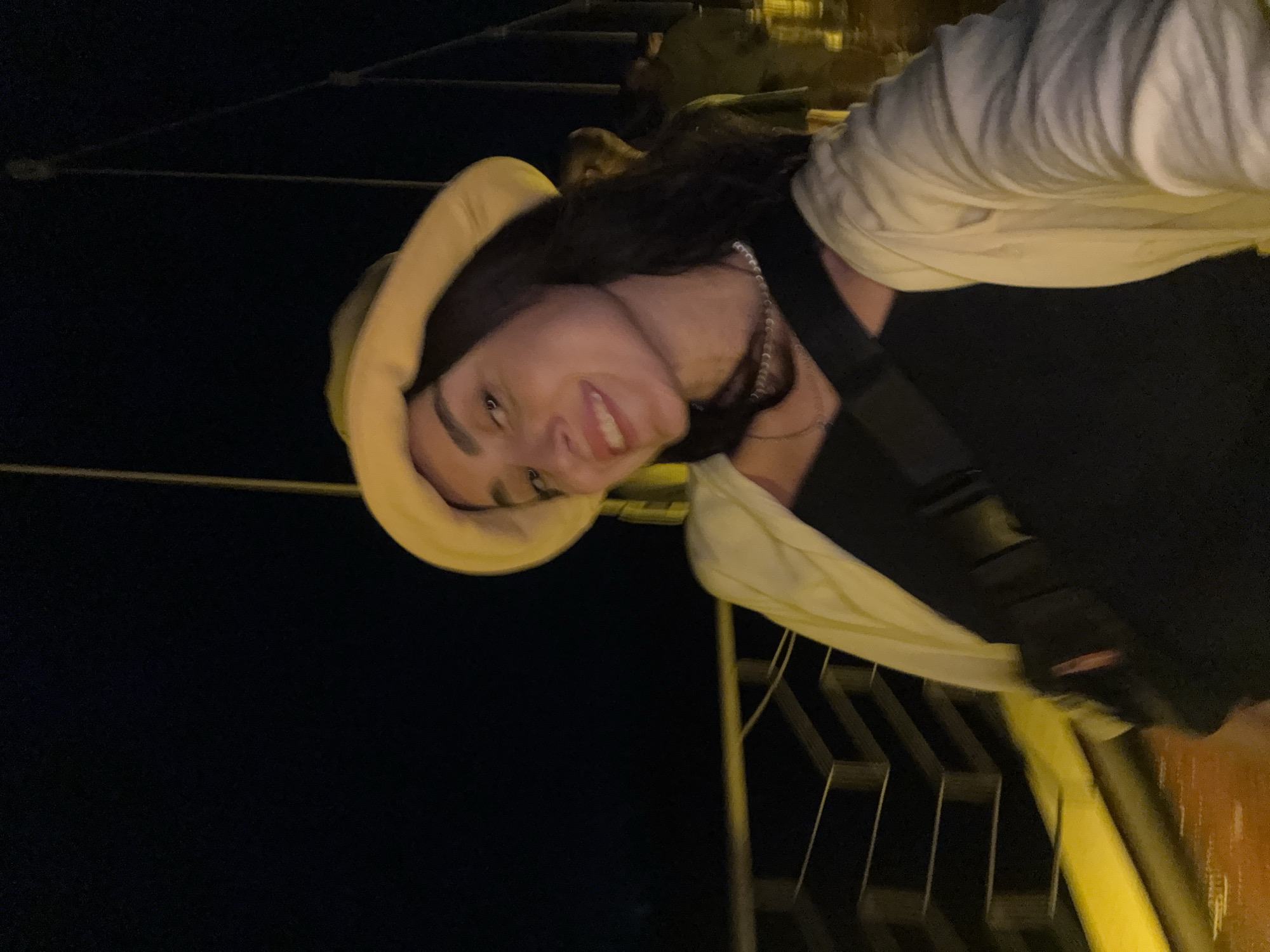 A young woman with dark hair and a light-colored bucket hat is looking at the camera and smiling. She is wearing a black top and a light-colored open jacket. A dark cross-body bag is visible over her shoulder. She is standing on a bridge with metal railings and yellow accents. The background is dark, suggesting it is nighttime. In the background, two figures are blurred and out of focus, also on the bridge. The bridge appears to be located in Poti, Georgia.
