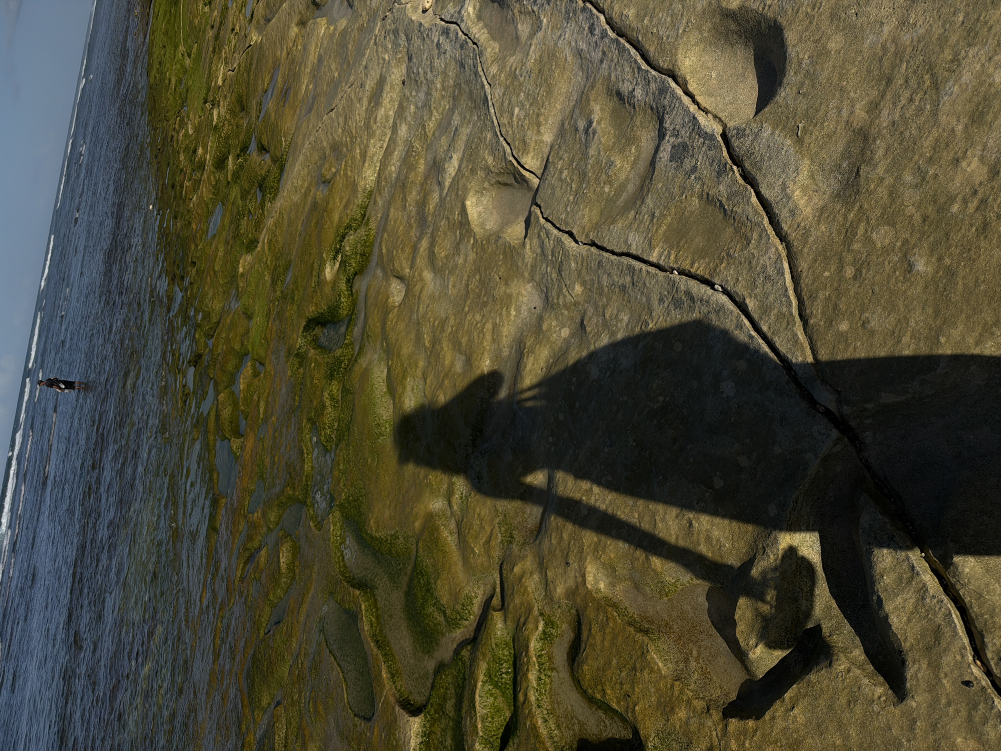 An eye-level, landscape-oriented, medium shot shows a shadow of a person on rock formations near the ocean at Kuta Selatan, Indonesia. The shadow stretches from the center of the image to the right. The person is standing on the left. The rock formations have cracks and some green algae. A person wearing dark clothes is in the water to the left. Waves wash onto the beach on the left side of the image. The sky is pale at the top left.