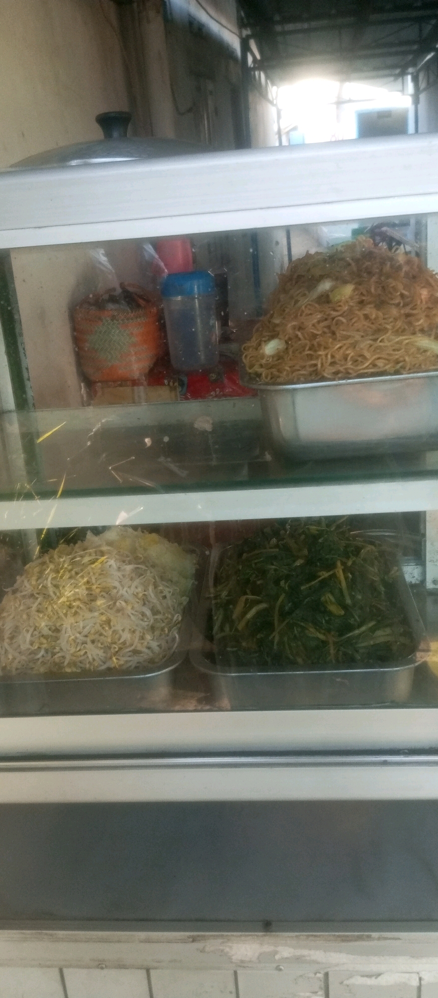 This image shows a food display cabinet, likely at a street food stall or small eatery, in Kediri City, Indonesia. The cabinet has multiple shelves filled with food items. The top shelf displays a large pile of stir-fried noodles and a woven orange basket containing what appears to be fried shallots or a similar garnish, next to a blue plastic container. The shelf below contains two metal trays, one filled with a mound of bean sprouts and the other with a portion of cooked green vegetables, possibly water spinach or a similar leafy green. The cabinet is made of metal and glass. The background is out of focus but suggests an outdoor or semi-outdoor environment, possibly a market or roadside. The overall impression is of a typical Indonesian food stall.