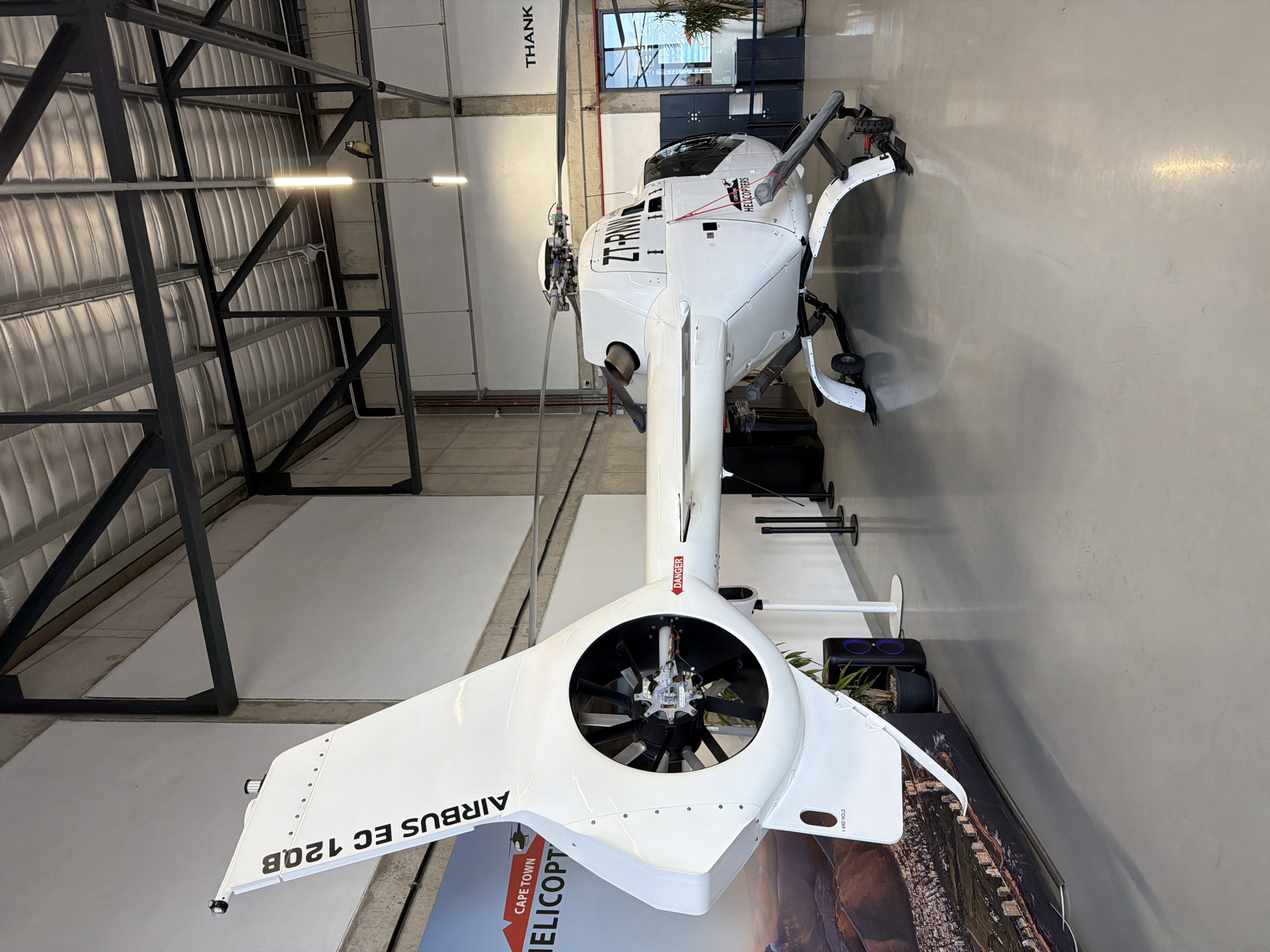 A white Airbus EC 120B helicopter, registered as ZT-RNN, is parked indoors in what appears to be a hangar or exhibition space. The helicopter is positioned at a slight angle, with its tail rotor assembly and rear fuselage prominently displayed on the left side of the frame. The text "AIRBUS EC 120B" is visible on the tail fin. The main rotor blades are also visible. The helicopter has landing skids and wheels. A large banner featuring the text "CAPE TOWN HELICOPTERS" and an image of a cityscape with mountains in the background is displayed to the left of the helicopter. The floor is a light grey, polished concrete. The ceiling is a metallic, corrugated material supported by dark metal beams and trusses, with overhead lighting fixtures. The walls are a light concrete color. In the background, there is a doorway with glass panes, and to the right of that, a large potted plant. A speaker system and a white stand are visible near the tail of the helicopter. The overall environment suggests a modern, well-lit indoor facility.