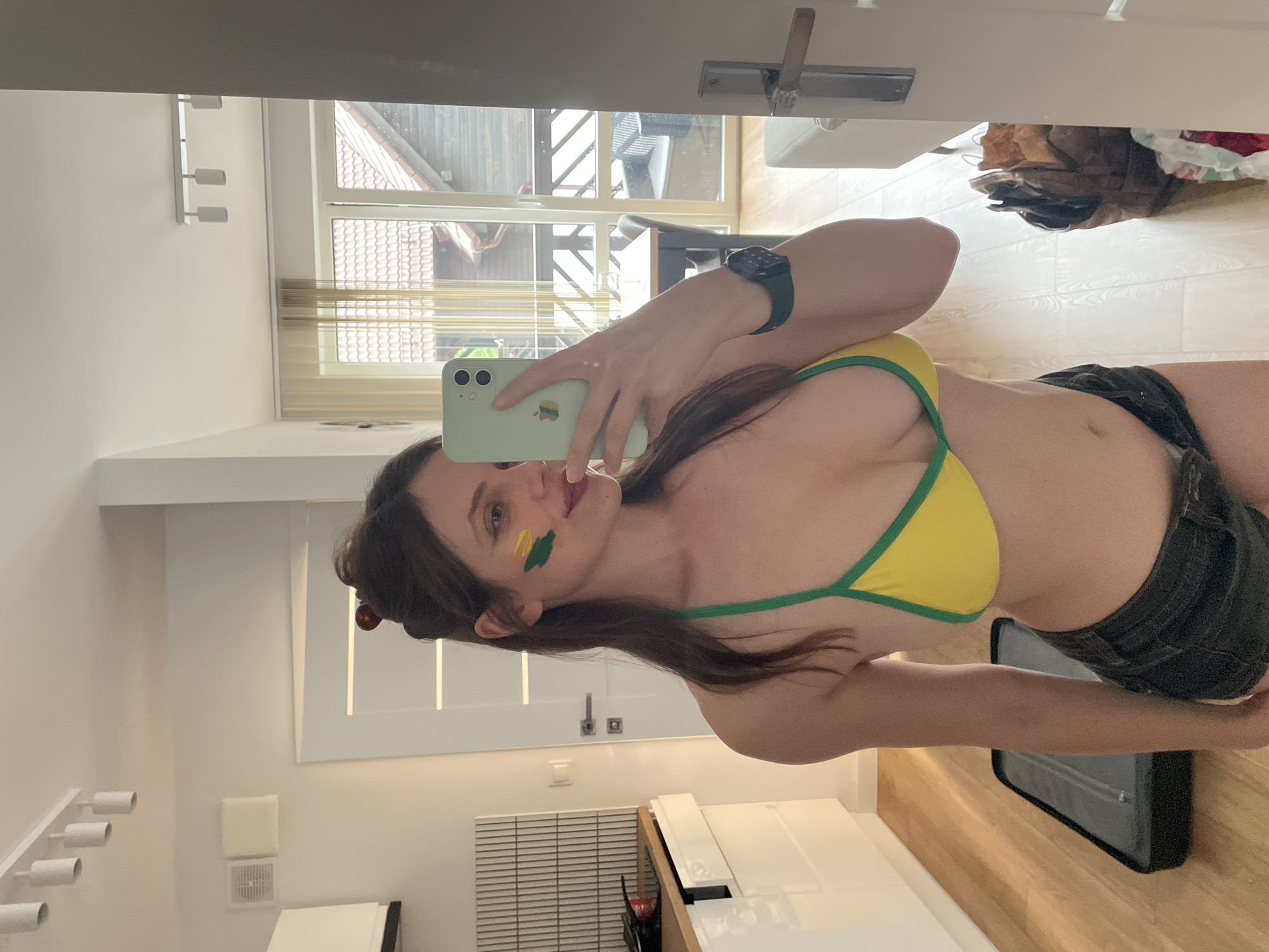 A young woman with brown hair styled in a bun takes a selfie in an apartment. She is wearing a yellow bikini top with green trim and dark denim shorts. She has green and yellow face paint on her left cheek. In her right hand, she holds a light green smartphone, with which she is taking the picture. On her left wrist, she wears a dark-colored watch.
The apartment appears modern and clean. To the left of the woman is a kitchen counter with a backsplash of black and white tiles. A white cabinet is beneath the counter. A black suitcase lies on the light-colored wooden floor to the woman's left. In the background, visible through a doorway, is a room with a table and chairs and a window overlooking a wooden building with a tiled roof. The view from the window suggests the location is in or near the city of Szklarska Poręba, Poland.