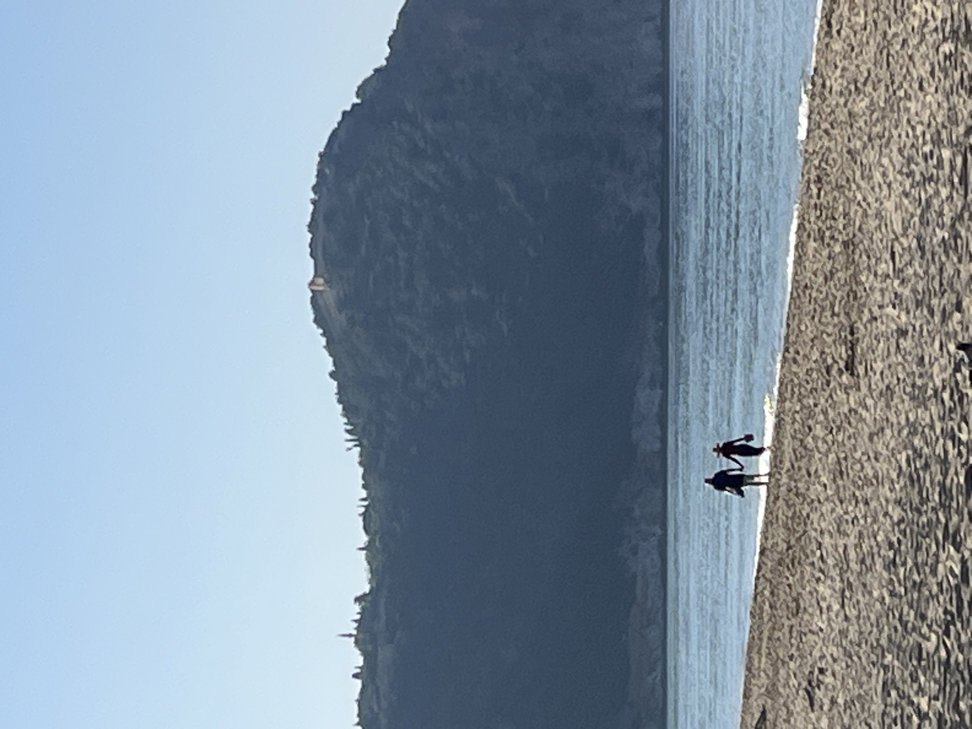The image shows a coastal scene with a mountain in the background and two people walking on the beach. The sky is clear and light blue. The water appears calm. The beach is made of coarse sand or pebbles. The mountain is dark and covered in vegetation. The image is likely taken in Alanya, Turkey, given the presence of the mountain and the coastal setting. The two people are silhouetted. They appear to be walking close together.