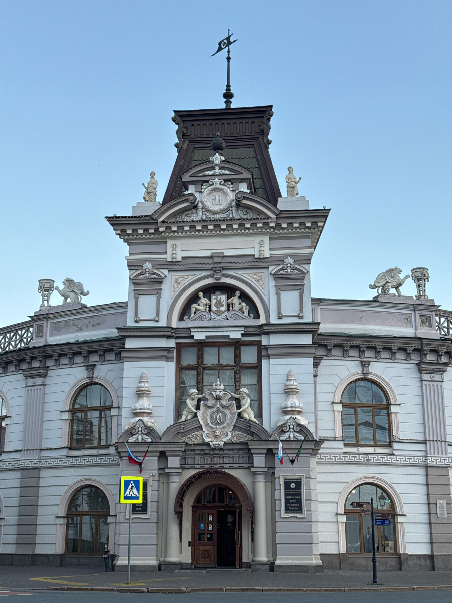 The image captures the facade of a historic building in Kazan, Russia, likely the National Museum of the Republic of Tatarstan. The building is ornate, featuring a central tower with a dark, mansard roof topped by a weather vane. Sculptural elements adorn the facade, including figures of women on pedestals, a winged lion-like creature on the left, and cherubic figures flanking a crest above the entrance. The building has a pale, grayish-blue exterior with decorative white trim and classical architectural details. Arched windows with dark frames are visible on multiple levels. In front of the building, a yellow pedestrian crossing sign is prominently displayed. Flags of Russia and Tatarstan are affixed to a pillar near the entrance. The sky is clear and pale blue, suggesting it is either early morning or late afternoon. The street in front of the building is paved, with a visible sidewalk and a trash receptacle.