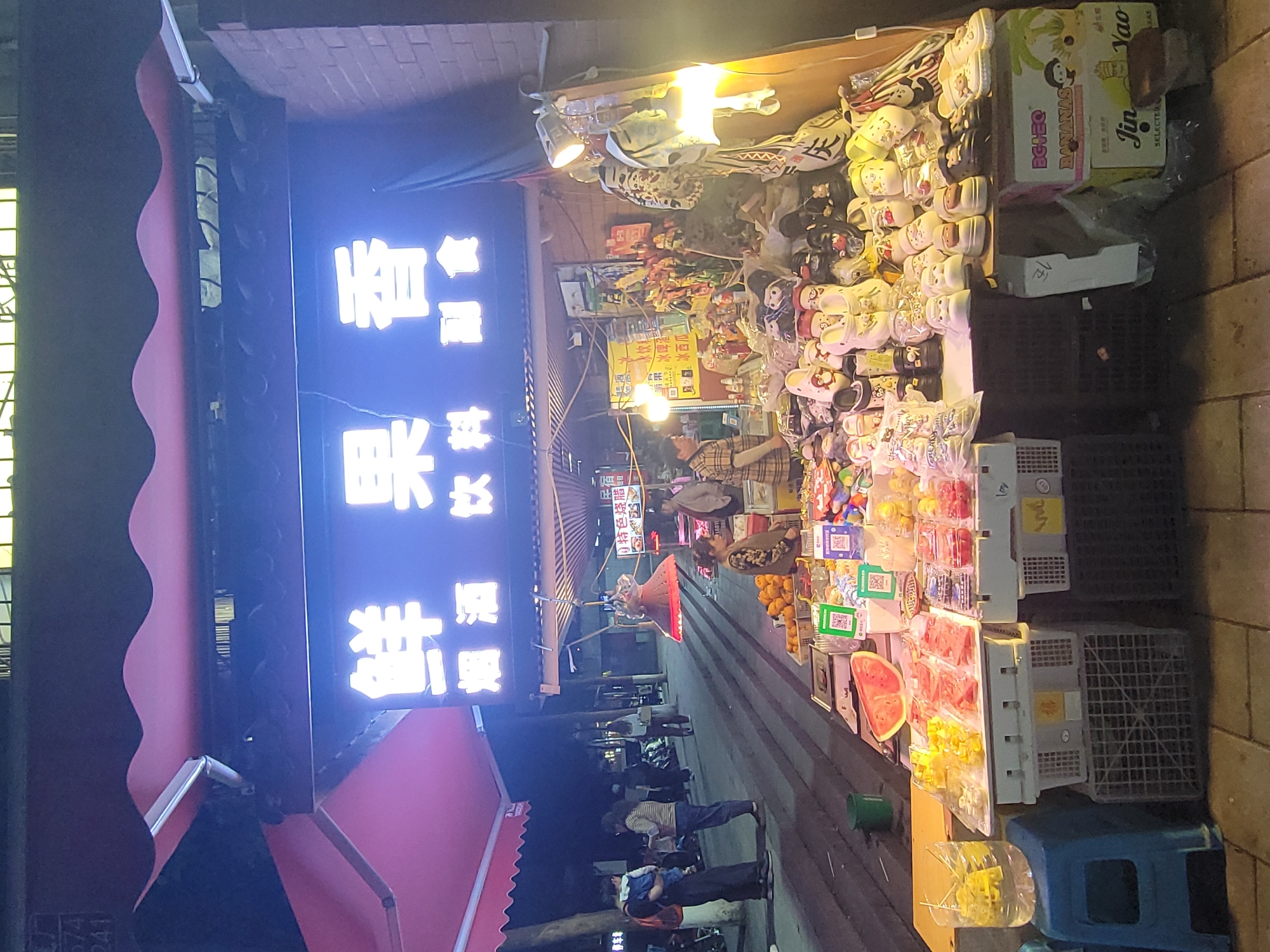 Here is a description of the image:
The image shows a street vendor stall in Yuzhong District, China, at night. The stall is overflowing with merchandise including fruit, drinks, packaged snacks, novelty items, and sandals. A blue illuminated sign above the stall displays Chinese characters. Several people are visible walking past the stall, browsing the merchandise, or standing around. The stall owner appears to be engaging with customers. The scene is lit by streetlights and the stall's own lighting. The ground is paved.