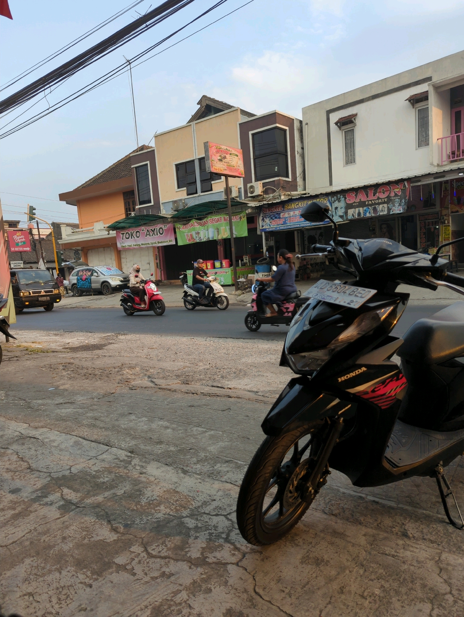 A street scene in Kacangan, Indonesia, features a black Honda scooter in the foreground, parked on a cracked concrete sidewalk. Behind the scooter, a street is occupied by various vehicles, including a black minivan, a silver car with stickers, a red scooter with a rider, and a white scooter with a rider. Several buildings line the street, displaying various shop signs, including "TOKO AXIO," "BANGKEL BANGKIT MOTOR," and "SALON FAVORIT." The sky is clear and blue, and the lighting suggests it is daytime.