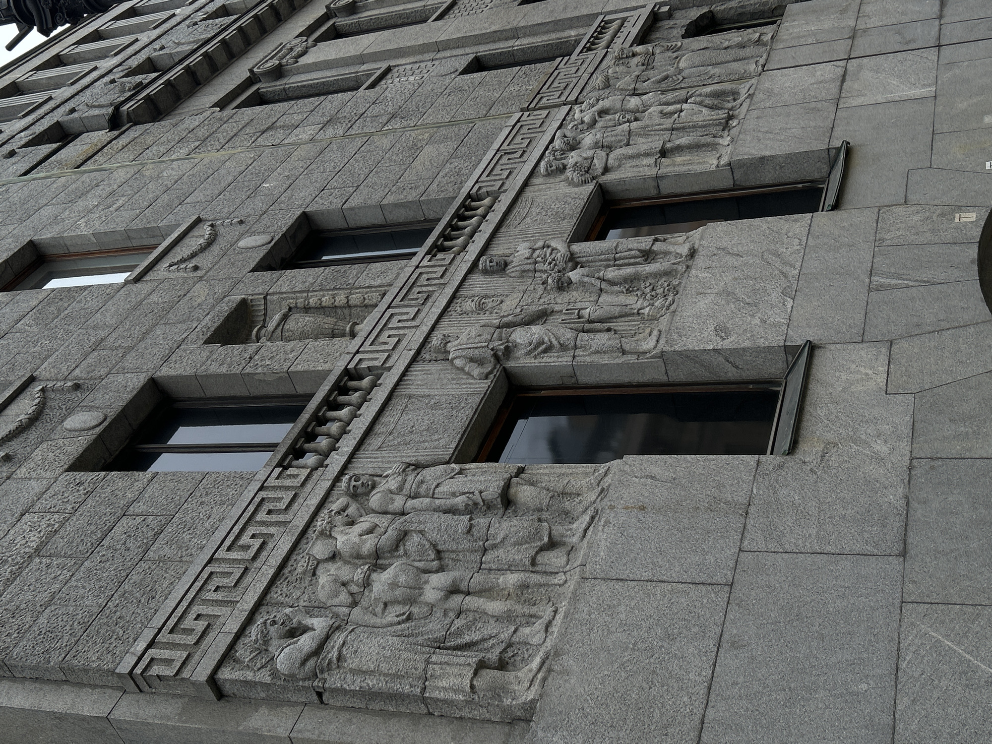The image depicts a close-up, low-angle view of a building facade. The building's exterior is constructed of gray stone blocks. The building is located in Saint Petersburg, Russia. The architecture exhibits detailed bas-relief carvings of human figures and decorative geometric patterns. Windows are set within the stone structure, appearing dark due to reflections or lack of interior light. A meander pattern is etched into the stone between the windows. The stone blocks show a uniform texture, and there are subtle variations in the color of the stone. The lighting is flat, suggesting an overcast day.