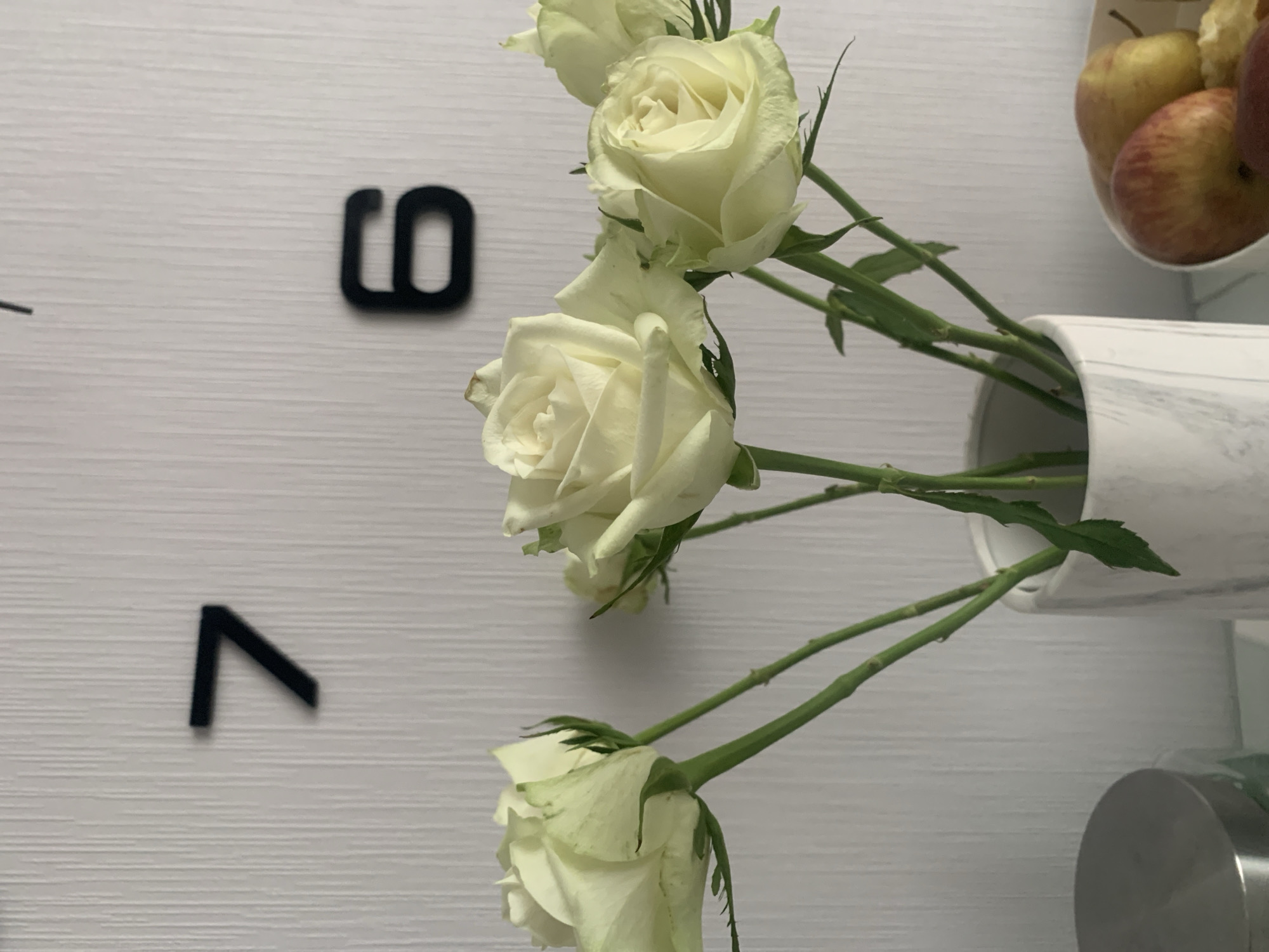 A close-up, low-angle shot shows a bouquet of pale green roses in a white marble vase. The roses have tightly packed petals with some light green tinges. Behind the roses, a white textured wall is visible, with two black numbers, a "7" and a "6," suggesting a clock face. To the right of the vase, a bowl of fruit, including apples and pears, is partially visible. In the lower left corner, the lid of a silver container can be seen. The overall lighting is soft and even. The scene is a still life composition, possibly set in a domestic environment in Kolpino, Russia.