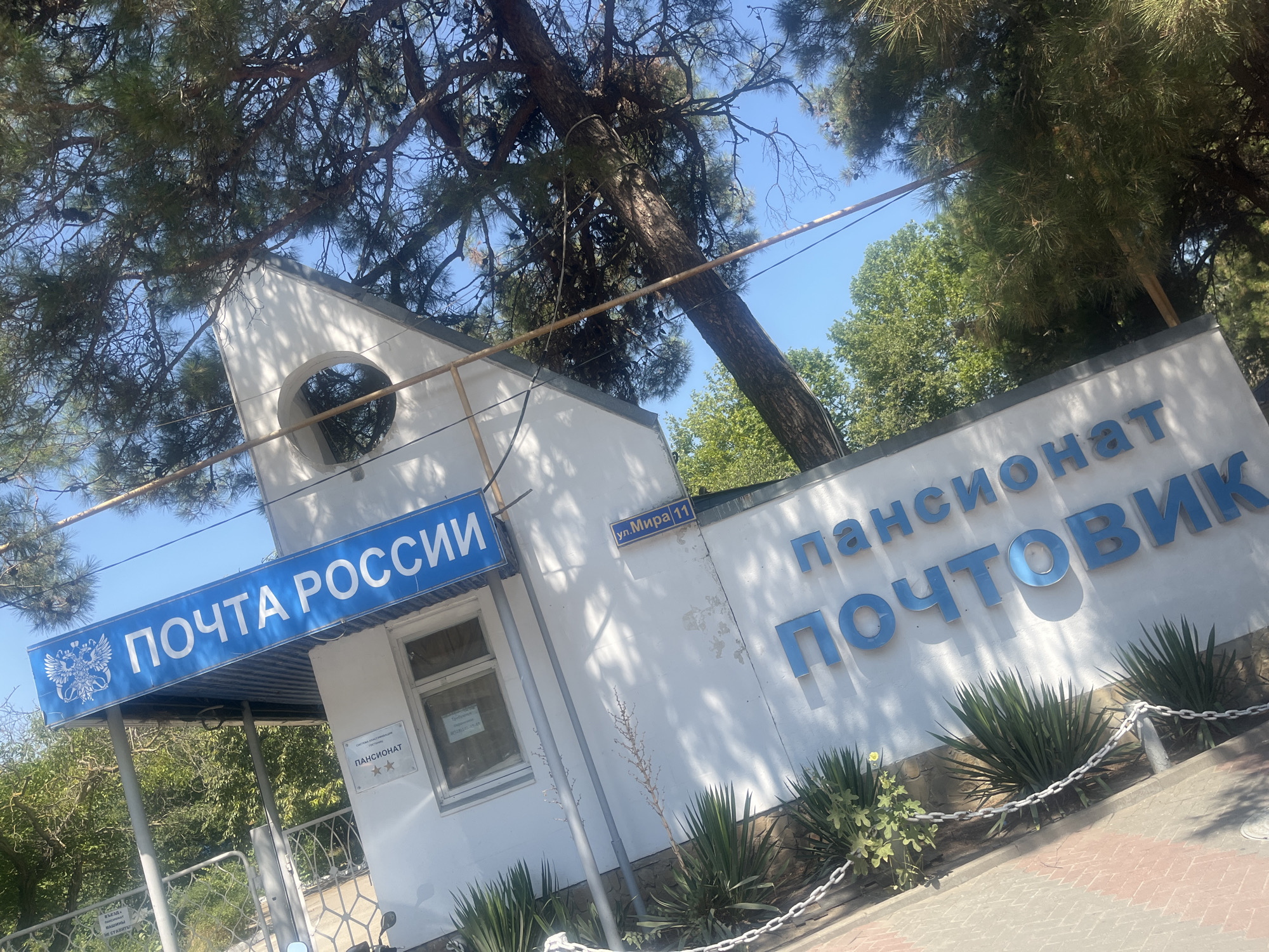 The image shows the building of the Pensionat "Pochtovik" in Novorossiysk, Russia. A blue sign with the inscription "ПОЧТА РОССИИ" (Post of Russia) and the emblem of the Russian Federation is mounted above the entrance. The building is white, with a triangular architectural element and a round window. The pensionat sign is in blue, block letters, with the inscription "ПАНСИОНАТ ПОЧТОВИК". There is also a street sign that reads "ул. Мира 11" (Mira Street 11). In front of the building, there are green bushes and a white chain. The sky is clear and blue, with branches of a large tree partially obscuring the view.