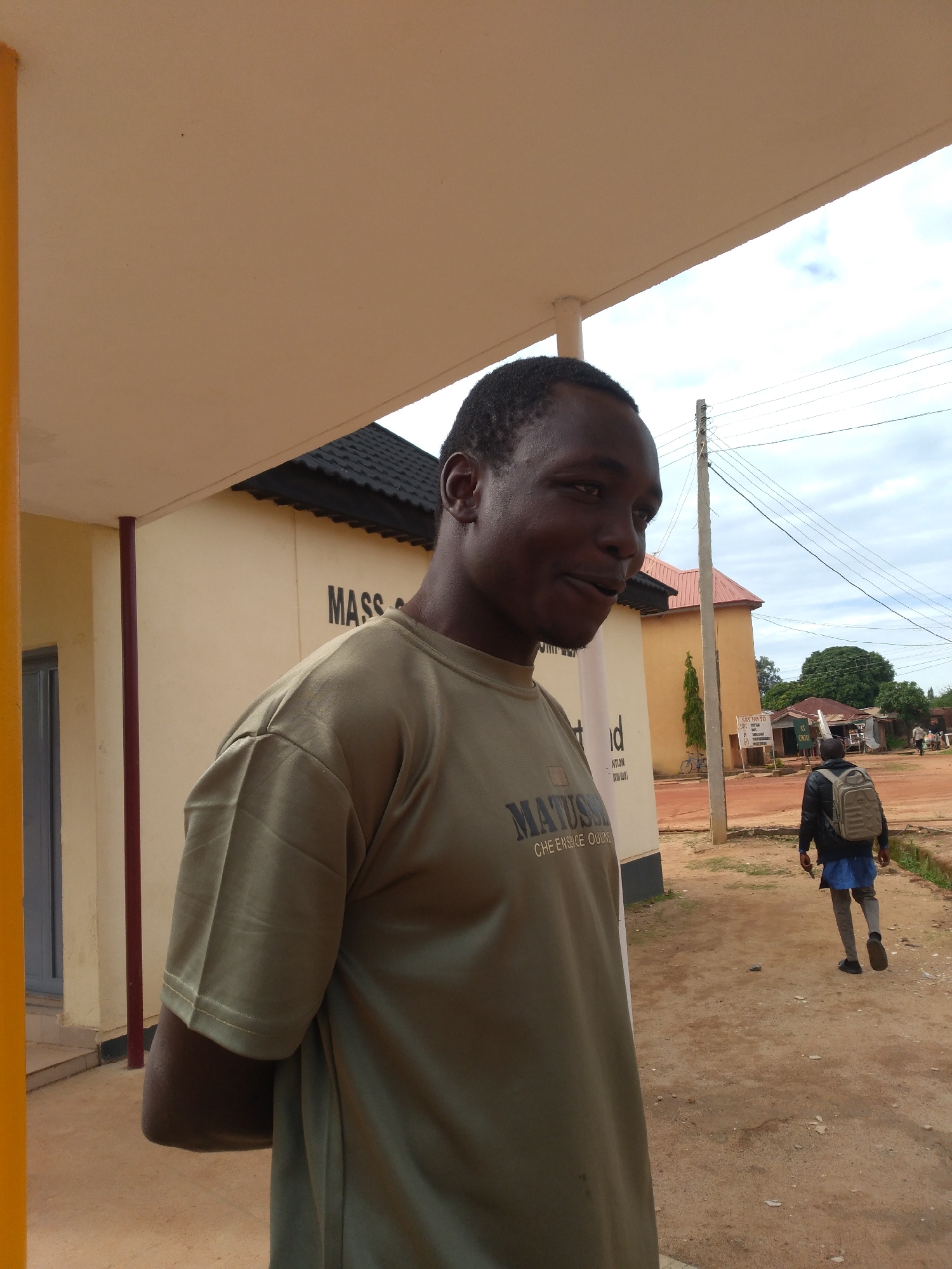 The image captures an outdoor scene in Jos, Nigeria, centered around a young adult male in the foreground. This male, of African descent, stands facing slightly to the right, looking out of frame with a subtle smile. He is wearing a light olive green short-sleeved t-shirt that displays partially visible dark lettering on the chest, reading "MATUSS" on the upper line and "CHE ENSUCE OULINE" on the lower line. His hands are held behind his back, mostly obscured. He stands under a light tan colored overhang or awning, which is supported by a prominent yellow vertical pillar on the far left and a narrower reddish-brown vertical support further to the right. The immediate ground beneath him is a light tan, possibly concrete, surface.
In the mid-ground, to the right of the primary subject, a light yellow building with a dark-tiled roof is visible, bearing the partial black text "MASS" on its side. A tall, light-colored utility pole with multiple overhead power lines extends vertically and horizontally across the right side of the frame. A second male figure, viewed from behind, walks away from the viewer on a reddish-brown dirt path or road. This individual wears a dark jacket, a blue garment underneath, light-colored trousers, and dark shoes, and carries a light beige or olive green backpack. Further in the background, additional low-rise buildings with various roof types and green trees are present. The sky above is overcast with visible white clouds.