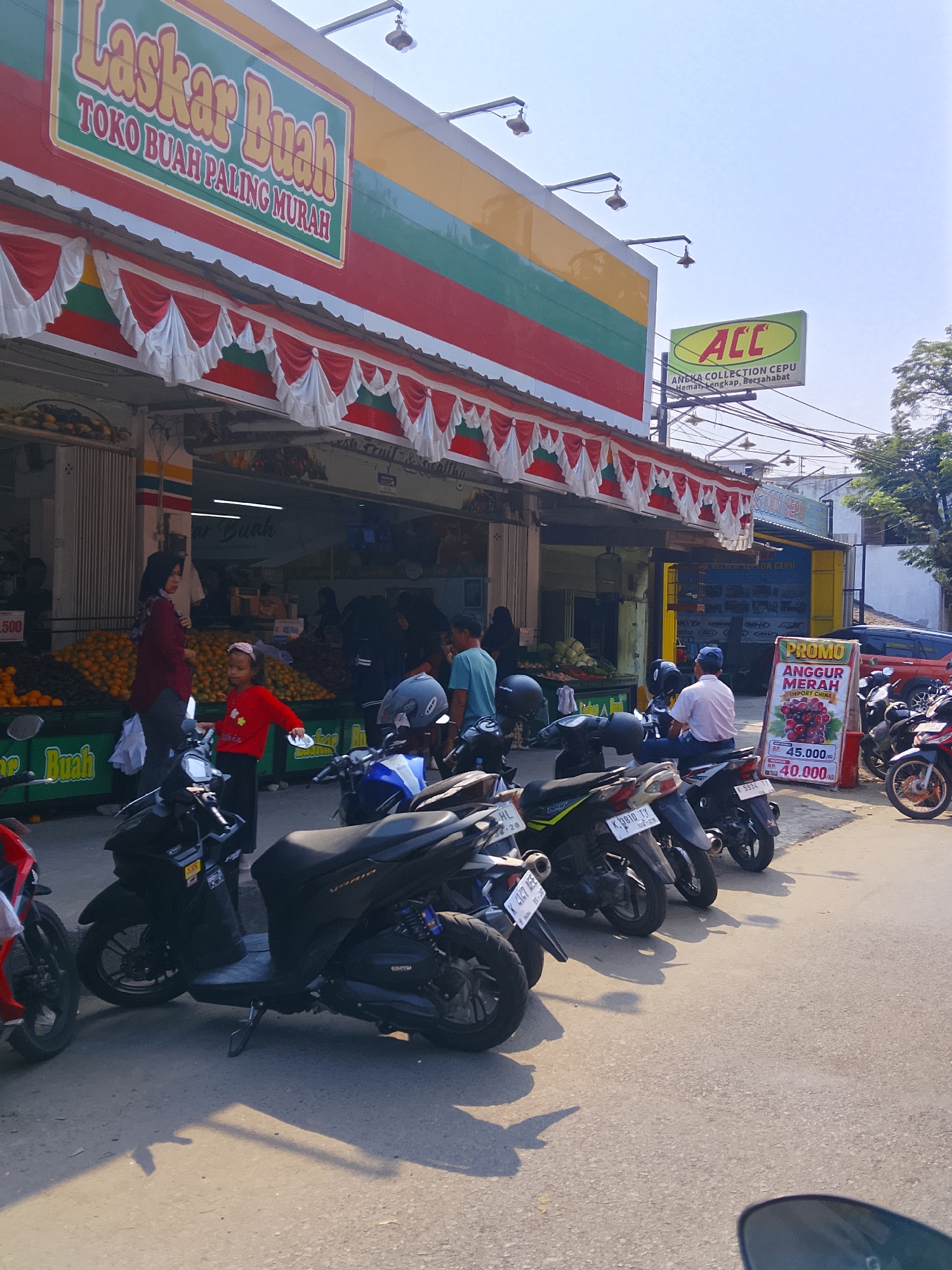 This is a medium shot, outdoor, street-level photograph taken in the city of Karangboyo, Indonesia. The scene depicts a busy street in front of a fruit market. A prominent sign above the market reads "LASKAR BUAH TOKO BUAH PALING MURAH" (Laskar Fruit Cheapest Fruit Store) in large, colorful letters. Another sign to the right advertises "ACC" and "ANEKA COLLECTION CEPU" (Various Collections Cepu).
In the foreground, several parked motorbikes line the street, indicating a common mode of transportation. A young girl wearing a red shirt and black pants is standing near a motorbike, looking towards the market. Behind her, a woman in a maroon headscarf and maroon top is interacting with someone inside the market.
The fruit market itself is visible behind them, with large displays of oranges and other produce. People can be seen browsing and shopping inside the market. The street is paved with asphalt and shows some wear and tear. The overall impression is of a vibrant, local marketplace on a sunny day.