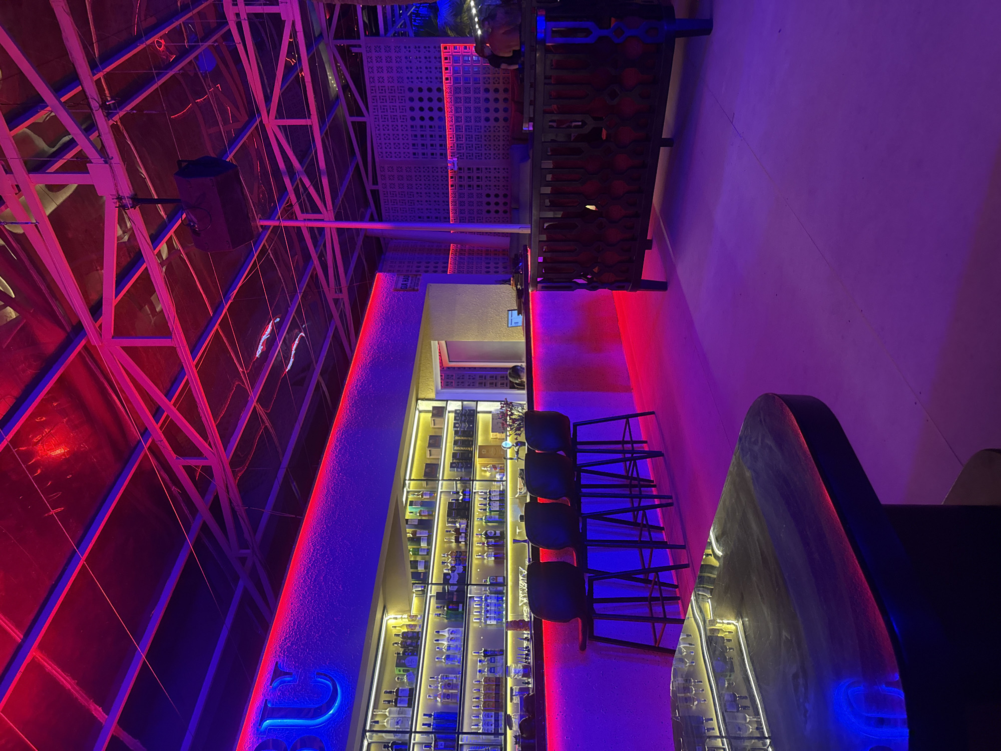 The image shows the interior of a bar or lounge in Unknown, Unknown. The architecture features an open, industrial-style ceiling with exposed metal beams illuminated by red neon lighting. A well-stocked bar with shelves of bottles runs along a wall, backlighting adding to the ambiance. Dark bar stools are placed along the bar. The lighting scheme includes blue and purple neon accents, creating a vibrant, modern atmosphere. A bar counter with reflective surfaces is visible in the foreground. No people are clearly visible in the foreground, but figures can be seen in the background.