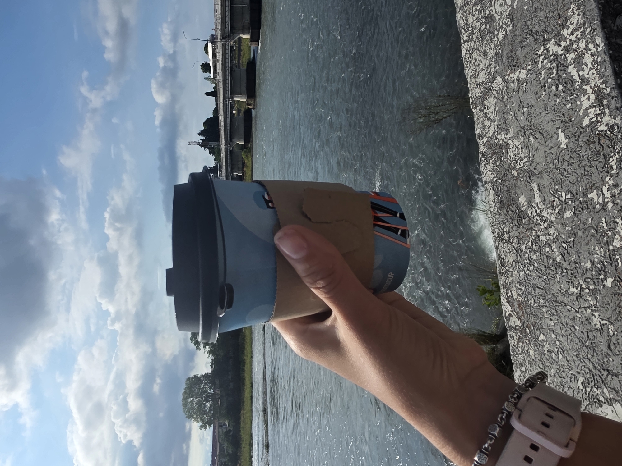 A hand holds a blue and white disposable coffee cup with a cardboard sleeve in front of a wide, choppy river under a cloudy sky. A bridge with concrete supports and a road carrying vehicles spans the river in the background. The scene appears to be outdoors, possibly near a promenade or waterfront, with a textured stone barrier in the foreground. The location is implied to be Sochi, Russia.