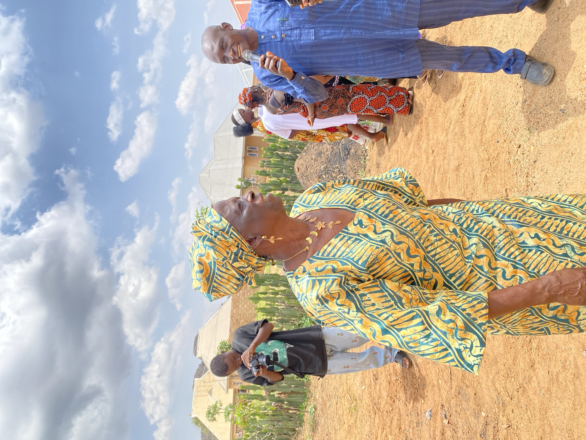 The image shows a group of people in Kuru Karama, Nigeria, on a dirt ground with some vegetation. In the foreground, an older woman in a yellow and green patterned outfit is looking up and to the left. Behind her, a man in a black shirt and grey pants is taking photos. Further back, a man in a blue striped shirt is holding a microphone and speaking. Behind him are two women in patterned dresses. Buildings are visible in the background, and the sky is cloudy.