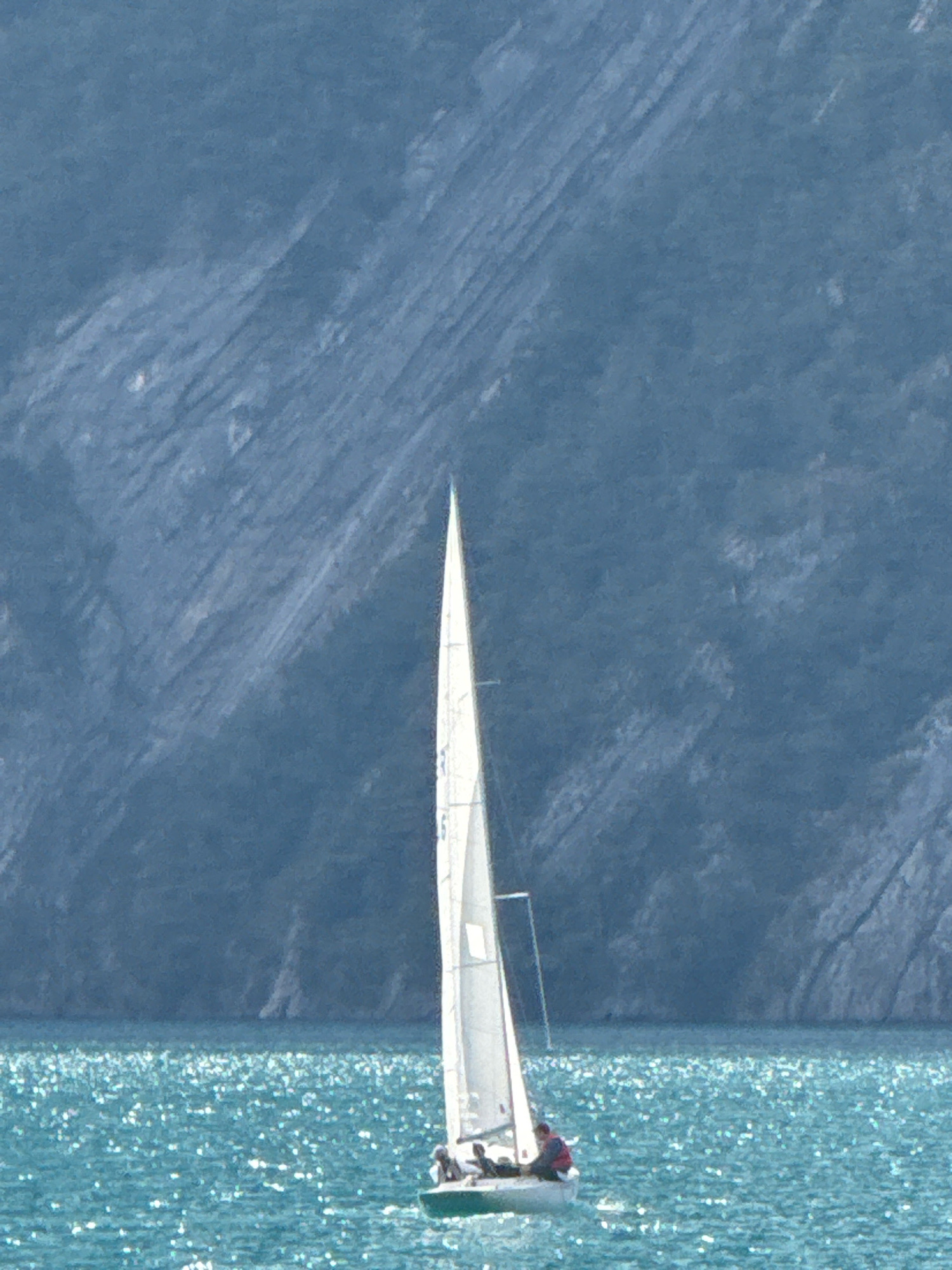 A sailboat with at least three people onboard sails on a body of water that sparkles with sunlight. The sailboat has a white sail and a green hull. Behind the sailboat, a steep, blue-grey mountain with visible striations rises sharply from the water. The sky above is not visible in the frame. The location is the lake near Sisikon, Switzerland.