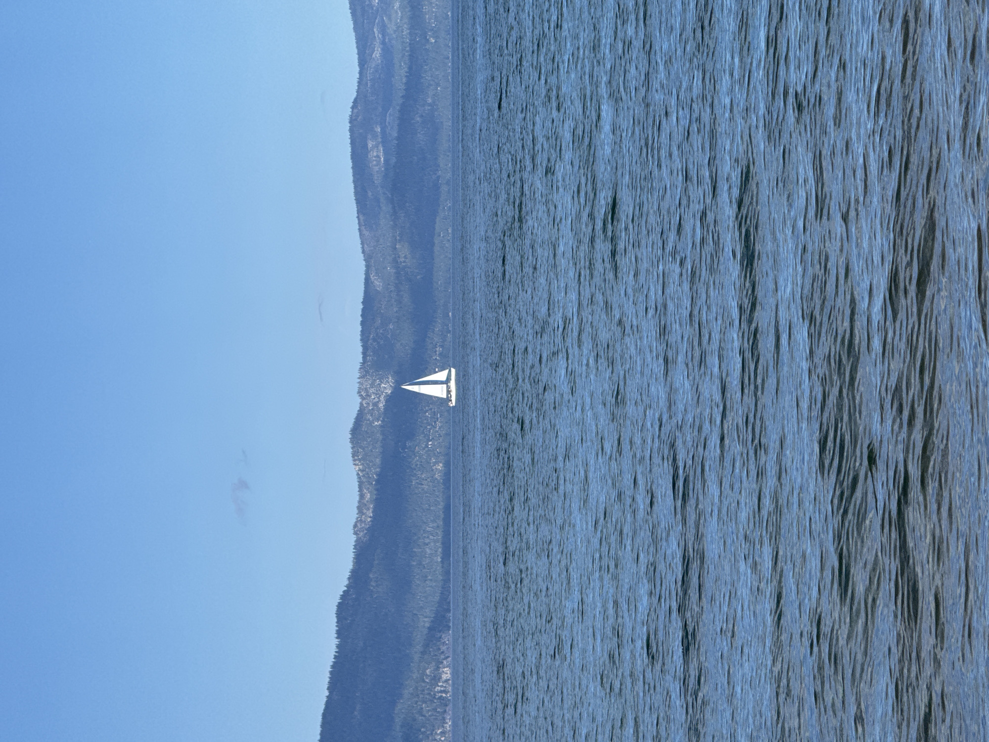 A lone white sailboat with two sails navigates across a calm, blue body of water. The water is textured with gentle ripples and reflects the clear blue sky. In the background, a range of dark, tree-covered mountains rises from the distant shore. The sky is a vast expanse of clear blue, with a faint wispy cloud visible in the upper left quadrant. The scene depicts a serene day on the water.