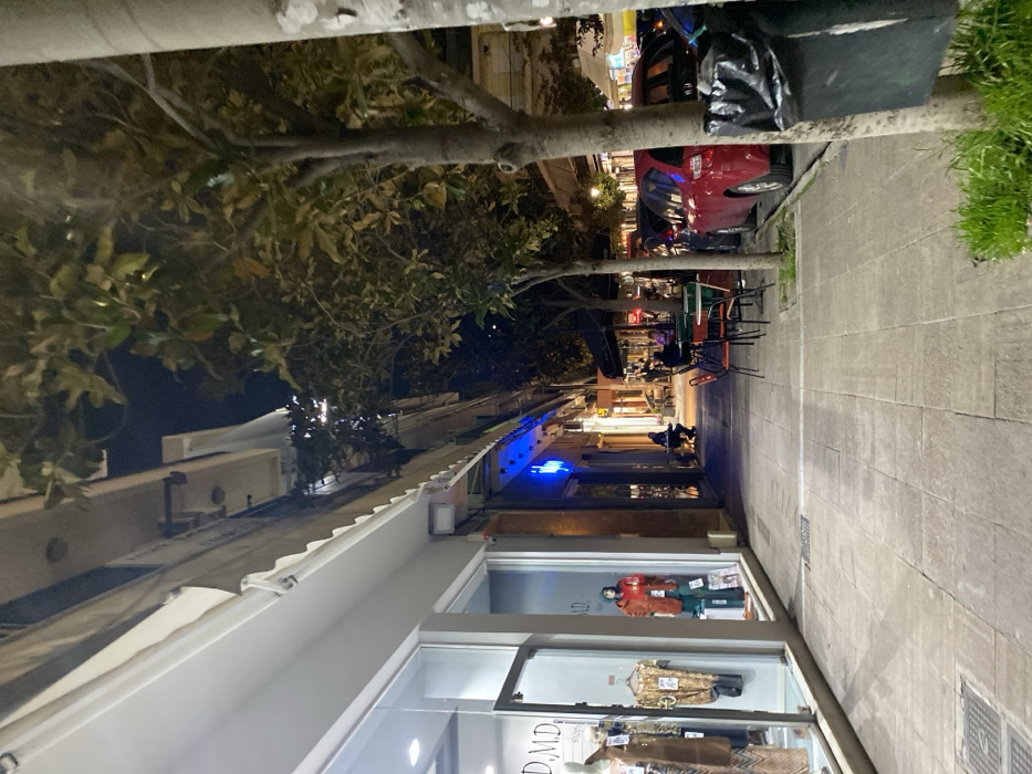 The image displays an evening street scene in Egaleo, Greece, looking down a commercial street at night. The sky is dark, with the scene illuminated by artificial lights.
On the right side, a paved sidewalk runs alongside buildings with shops. In the foreground, a shop window features mannequins displaying clothing items. The text "D.M.D signa" is visible on this window, likely part of a brand name. A light fixture is mounted on the white awning above the shop.
Trees with substantial trunks and leafy branches line the street to the left. Further down the street, a red car is parked, and a black trash bag is tied around the base of one of the tree trunks nearby. Beyond the parked car, more businesses are visible, characterized by illuminated storefronts and awnings. Outdoor seating with tables and chairs suggests cafes or restaurants, where a few blurry figures of people can be seen. A bright blue neon sign stands out on one of the buildings in the distance. The overall impression is that of a lively urban environment during evening hours.