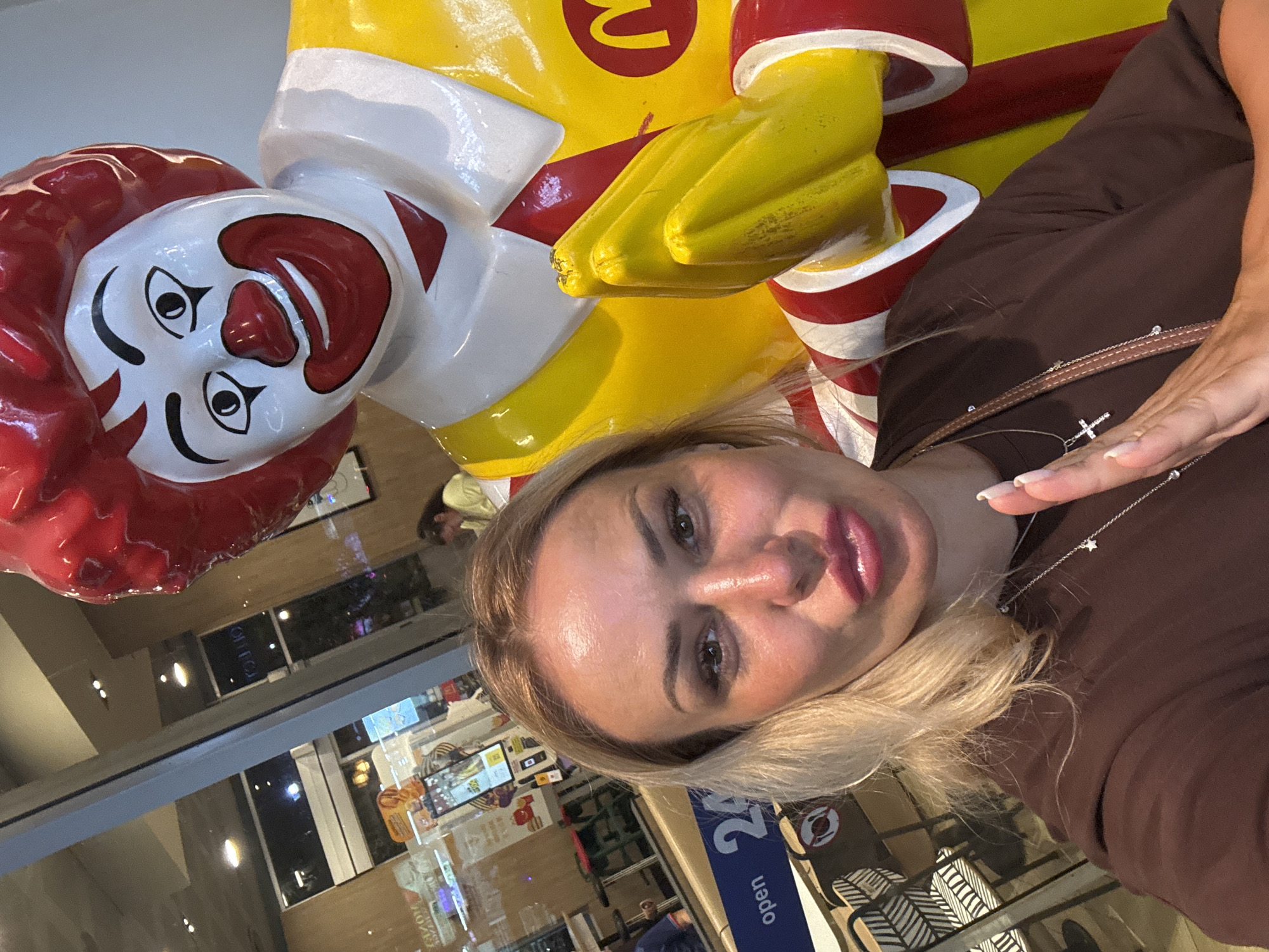A woman with blonde hair and red lipstick takes a selfie in front of a Ronald McDonald statue. The woman is wearing a brown t-shirt and a silver necklace with a cross pendant. The Ronald McDonald statue is large and colorful, with a red wig, white face paint, and a yellow and red uniform. In the background, a McDonald's restaurant is visible through a glass window, with signs indicating "open 24". Other patrons can be seen inside the restaurant. The scene appears to be a public space within a city.