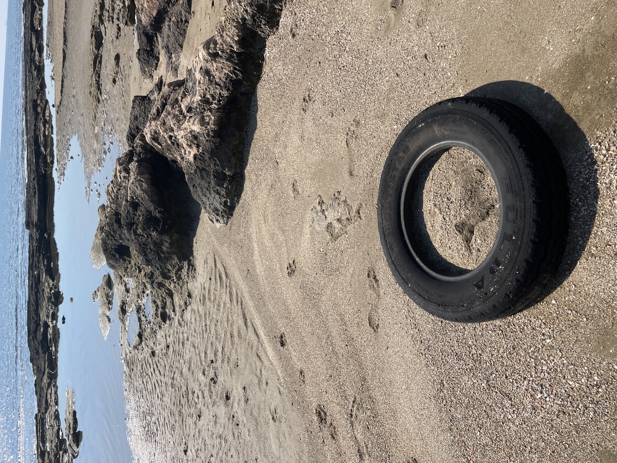 An abandoned car tire lies in the sand on a rocky beach near the ocean in Spathariko, Northern Cyprus. The tire is black and circular, with some text visible on its side. The beach is composed of sand, small shells, and scattered rocks. In the background, the calm blue ocean stretches to the horizon, with the sun reflecting off its surface. There are tide pools and rocky outcrops visible along the shoreline. The scene is quiet and appears deserted, with no people present.