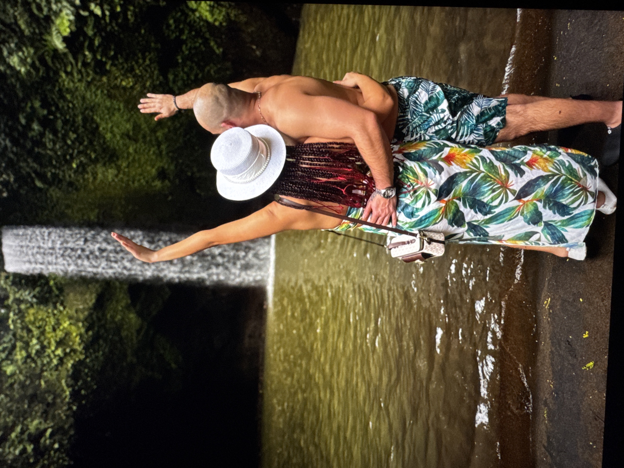 A man and woman stand facing a waterfall. The man is shirtless and wearing a dark green and white patterned bathing suit. The woman is wearing a long, white floral-patterned dress and a white hat. They both have their arms outstretched. The location is likely a natural setting, and the city and country are unknown.