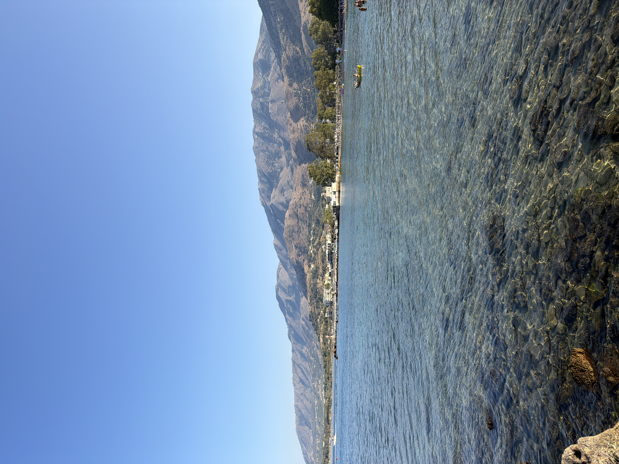 The image shows a view of the clear, shallow waters of Georgioupoli, Greece, with the coastline and mountains in the background. The water is transparent, revealing a rocky seabed. Several people are visible in the water, some swimming and others in a yellow kayak. Buildings and trees line the shore, and a lighthouse can be seen in the distance. The sky is clear and blue.