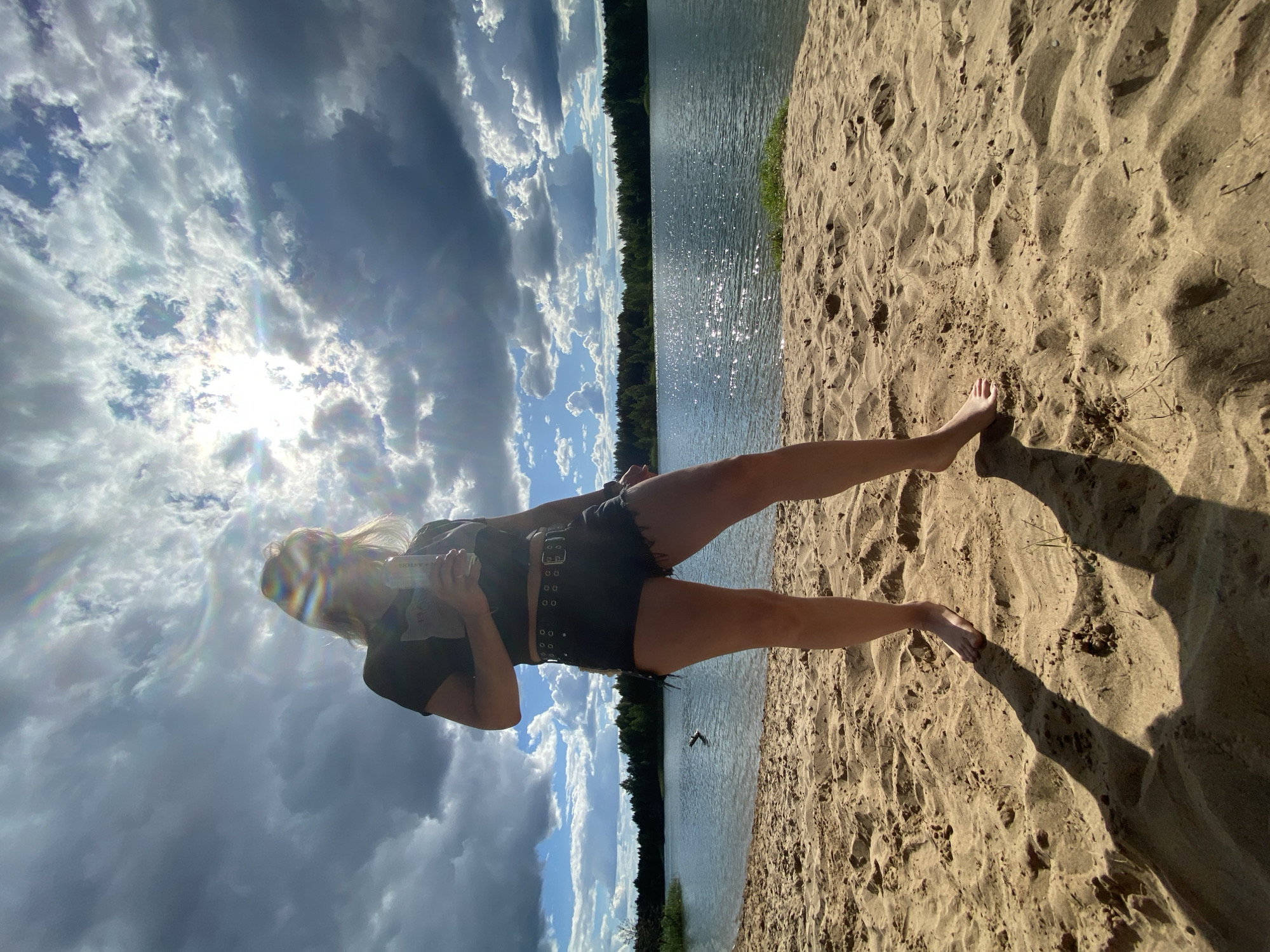 A person stands on a sandy beach, holding a can. The person is wearing a black crop top and black denim shorts. The background shows a body of water and a treeline under a dramatic sky with clouds and sunlight. The location is the beach of Lake Bely, Novyi, Russia.
